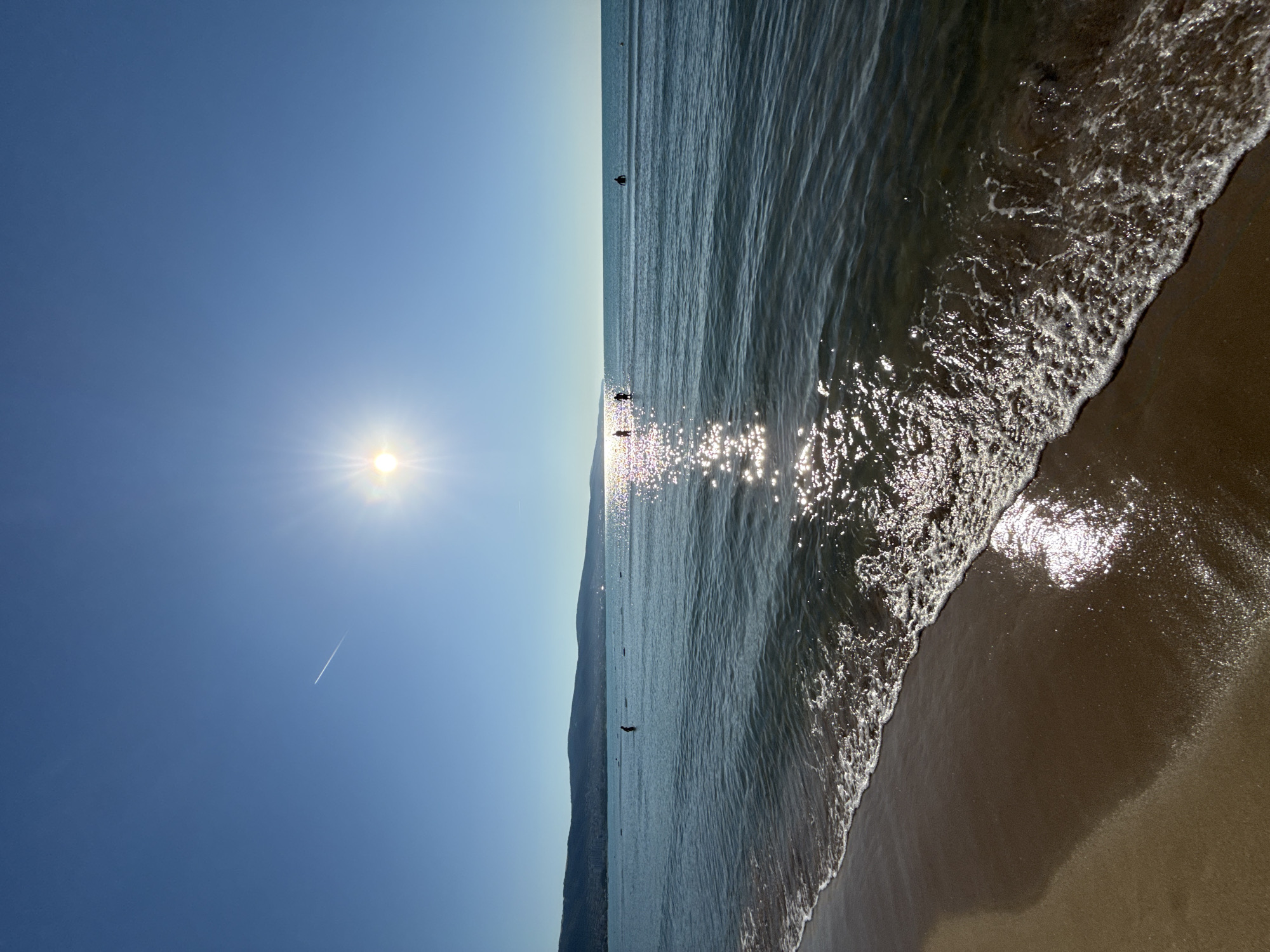 An outdoor, eye-level, full shot captures a beach scene on a sunny day in Nesebar, Bulgaria. The sun shines brightly in the upper quadrant of the frame, casting a dazzling reflection across the calm sea. A single, white contrail from an airplane arcs across the clear blue sky. The foreground shows a sandy beach with gentle waves lapping at the shore. In the middle ground, several people are visible in the water; one individual stands alone near the shore, while others are further out, appearing to swim or wade. In the background, a distant coastline with a ridge of hills topped with buildings stretches across the horizon. The water is a deep blue, transitioning to lighter shades near the shore where it is clearer and the sandy bottom is visible. The overall impression is one of a peaceful and serene coastal environment.