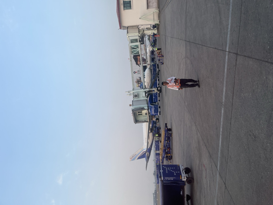 The image captures an airport tarmac scene, likely depicting ground operations around a parked aircraft. A clear blue sky dominates the upper left, transitioning to a lighter, somewhat hazy blue towards the horizon on the right.
The central focus is a male individual, appearing to be airport ground staff, standing on the concrete apron. He wears a bright orange safety vest over a white shirt, dark trousers, and dark shoes. In his left hand, he holds a small, white rectangular object, possibly a manifest or electronic device. He is facing slightly towards the left of the frame.
Behind him, an IndiGo airline aircraft, distinguished by its blue and white livery, is parked at a gate. Its tail fin, engine, and a portion of the fuselage are clearly visible. A jet bridge is connected to the aircraft, suggesting boarding or de-boarding operations are in progress or recently completed. Further behind this plane, the nose section of another aircraft is partially visible.
Numerous pieces of airport ground support equipment are scattered across the tarmac. Several blue baggage carts or loaders, some explicitly branded with "IndiGo" and text like "SUPER CAPACITY / 1.7 M³", are positioned near the aircraft, indicating baggage handling activities. A building, likely part of the airport terminal, stands in the background, with windows and a white and beige exterior. The concrete ground features white demarcation lines.
The scene suggests a daytime setting with fair weather. The overall activity portrayed is the routine servicing and handling of an aircraft at an airport, specifically in Guwahati, India.