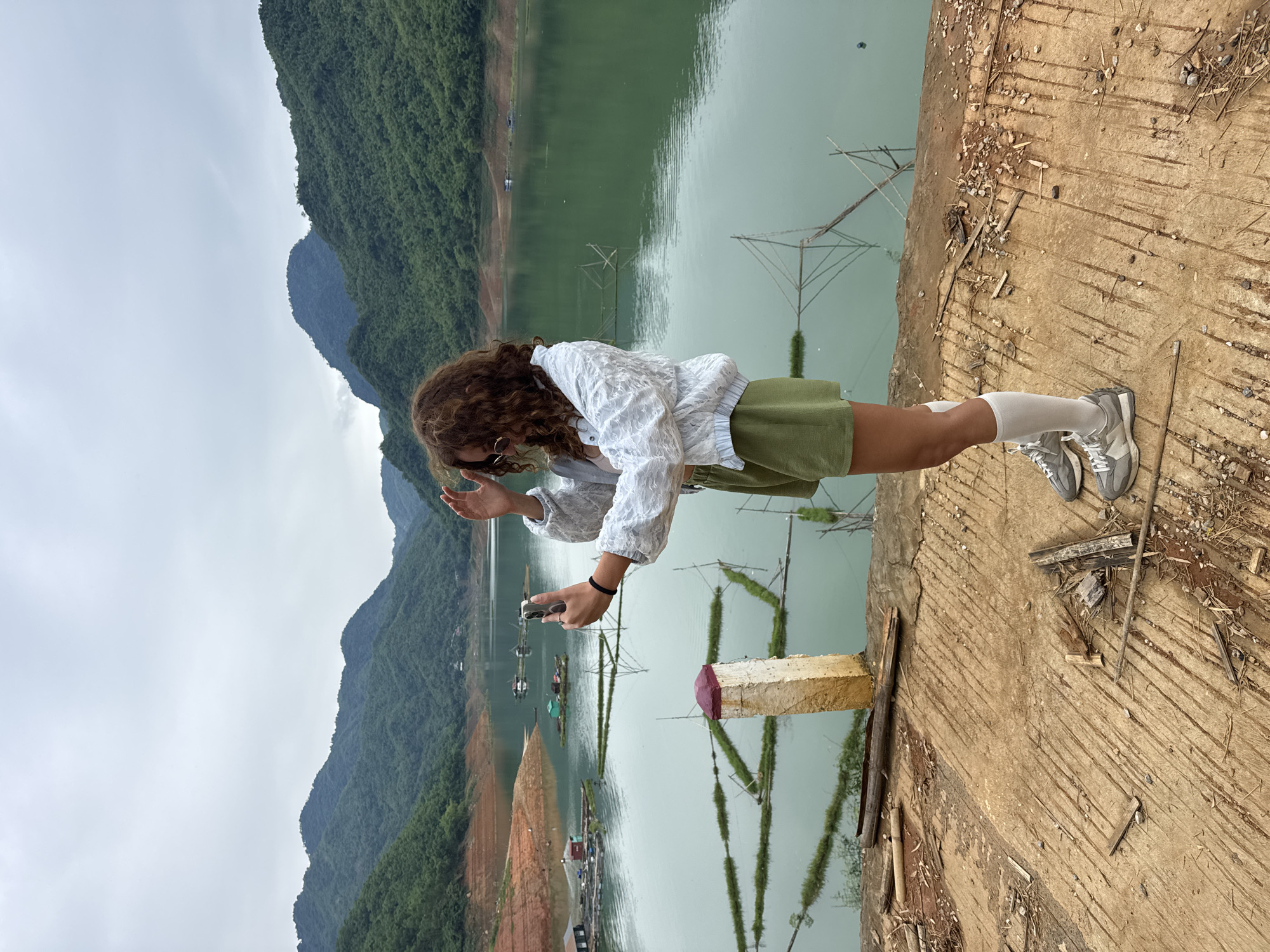 A young woman with curly brown hair is taking a selfie with her phone in front of a scenic view of a lake and green mountains. She is wearing a white textured jacket, olive green shorts, white socks, and gray sneakers. She is standing on a rough, textured concrete surface with debris scattered around her. The lake in the background is a greenish-blue color and appears to be a reservoir, with some floating structures and fishing nets visible. The mountains are lush and green, with some terraced areas visible on the slopes. The sky is overcast. The location is the reservoir at Bản Phúc, Vietnam.