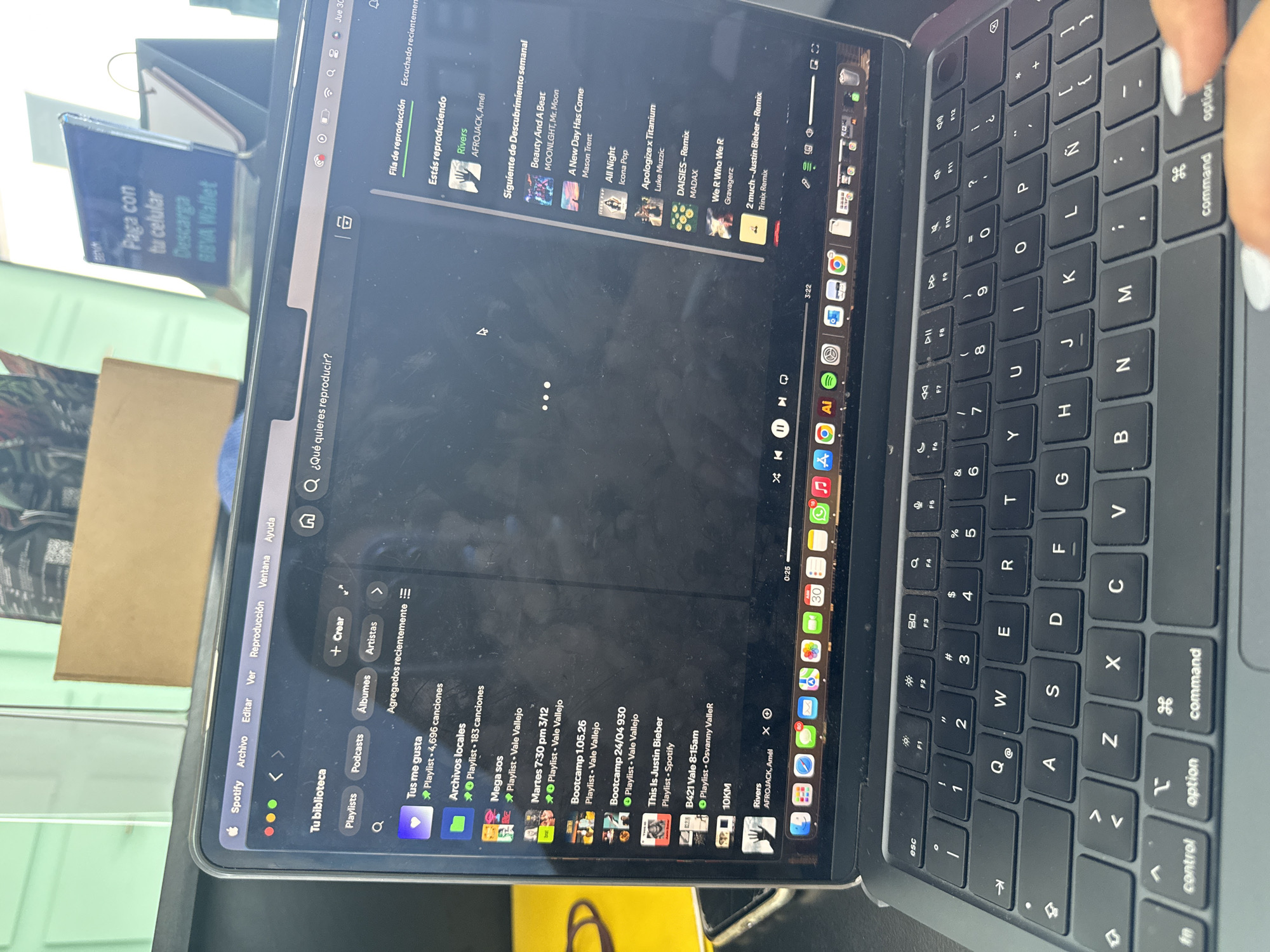 A high-angle, slightly rotated shot captures a person interacting with a laptop, likely a MacBook, which is open on a surface, possibly a desk, in an indoor setting. A human hand with white painted nails is visible on the Magic Keyboard, positioned near the "option" and "command" keys on the right side.
The laptop screen displays the Spotify application, showing both the music playback interface and the user's library. In the "Estás reproduciendo" (Currently playing) section, "Rivers" by AFROJACK, Amél is highlighted. The "Siguiente de Descubrimiento semanal" (Next from Weekly Discovery) queue lists several songs, including "Beauty And A Beat," "A New Day Has Come," "All Night," "Apologize x Titanium," "DAISIES - Remix," "We R Who We R," and "2 much - Justin Bieber - Remix."
The Spotify library on the left shows "Tus me gusta" (Your likes) with "4,696 canciones" (4,696 songs) and "Archivos locales" (Local files) with "183 canciones" (183 songs). Various playlists are listed, many attributed to "Vale Vallejo" and "Osvanny ValleR," such as "Mega sos," "Martes 7:30 pm 3/12," "Bootcamp 1.05.26," "Bootcamp 24/04 930," "This Is Justin Bieber," "B421 Vale 8:15am," and "10KM."
In the background, a phone in a stand is partially visible, displaying text in Spanish: "Paga con tu celular Descarga BBVA Wallet." The top right corner of the laptop screen shows "Jue 30" (Thursday 30) and the time "3:22." The screen appears somewhat smudged or dusty. The keyboard features a Spanish layout. The scene is likely in San Bernardino Tlaxcalancingo, Mexico.
