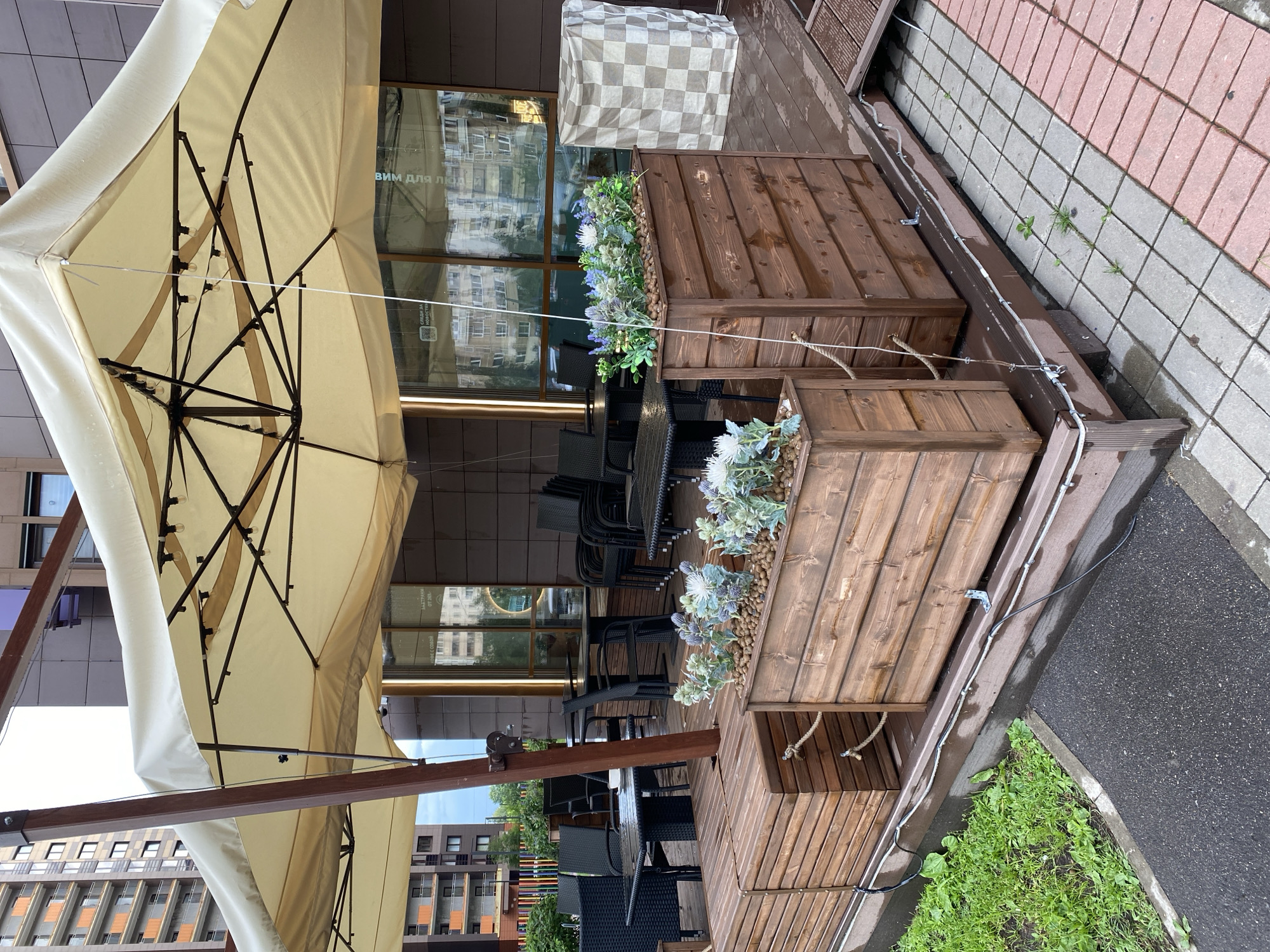 This image shows an outdoor seating area of a cafe or restaurant. A large cream-colored umbrella provides shade over several tables and chairs. The furniture appears to be made of dark wicker. In the foreground, there are wooden planter boxes filled with artificial flowers and pebbles. The planter boxes are integrated into a wooden platform. A checkered tarp is draped over a structure behind the planter boxes. The background shows the building facade with large windows reflecting the surrounding environment. The ground is paved with interlocking stones and asphalt. Some green grass grows along the edge of the paved area. The scene appears to be set during the day, possibly after rain, as there are some water droplets visible on the umbrella. There is no visible activity or people in the image. Text is visible on the windows in the background, but it is blurry and not legible.
