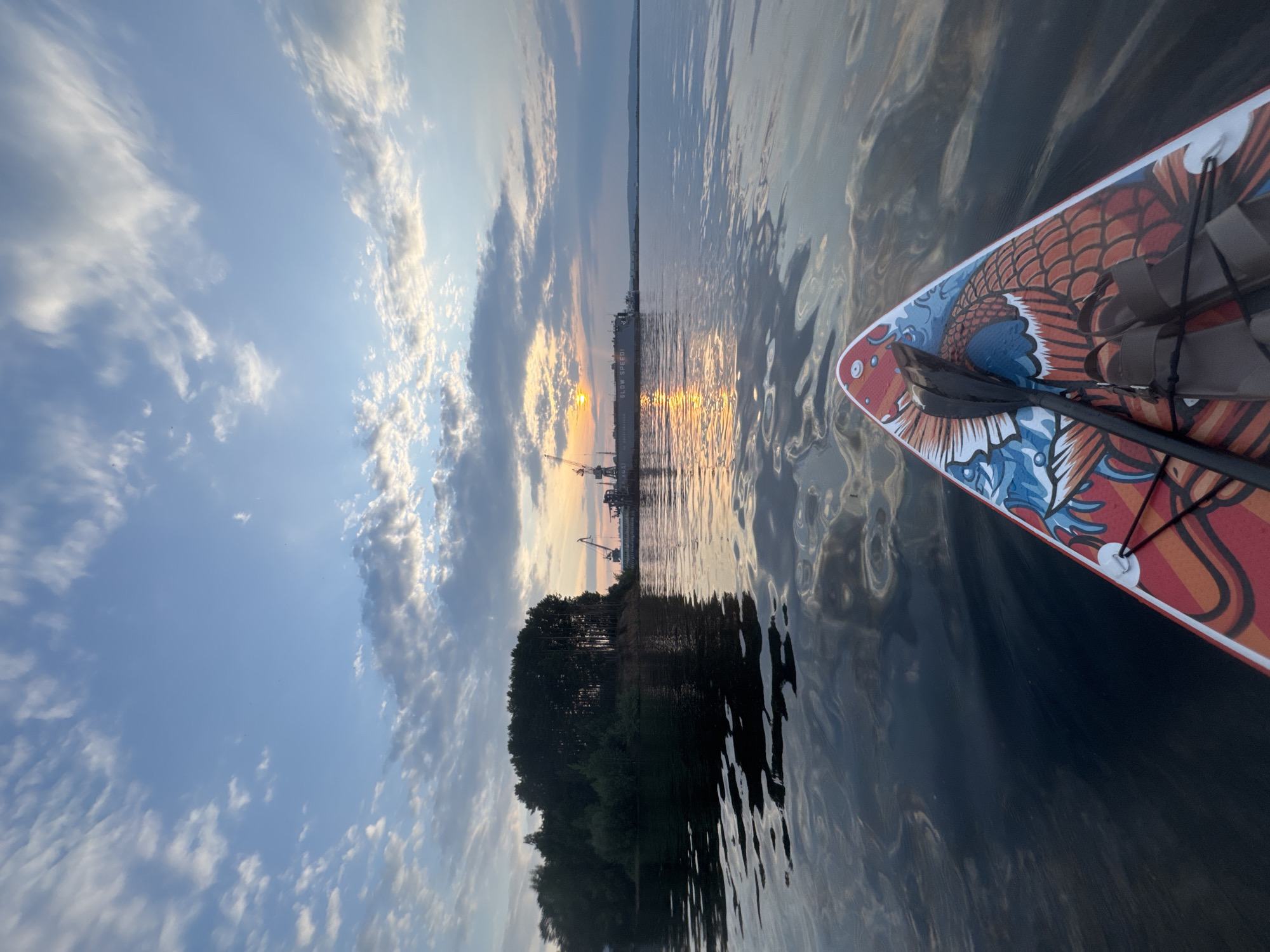The image depicts a first-person perspective from a paddleboard on a body of water, likely at sunset, with a scenic view of Chaikovsky, Russia. In the foreground, the tail end of a paddleboard is visible, adorned with a vibrant, artistic design featuring a phoenix or similar mythical bird motif in red, orange, and blue. A paddle rests on the board, and a pair of brown sandals are situated on the deck. The water is calm with ripples and reflections of the sky and distant objects.
The middle ground of the image shows a forested island on the left, with dense dark green trees lining its shore. To the right of the island, a large industrial structure is visible across the water, featuring cranes and what appears to be a docked ship or barge. The ship has the words "SLOW SPEED" written on its side. The setting sun casts a warm, golden glow on the water and the clouds in the sky.
The sky is a mix of blue with scattered white and gray clouds, illuminated by the setting sun. The overall atmosphere is tranquil and serene, capturing a moment of outdoor recreation against an industrial and natural backdrop. The location is identified as Chaikovsky, Russia, suggesting a waterfront area within or near the city.