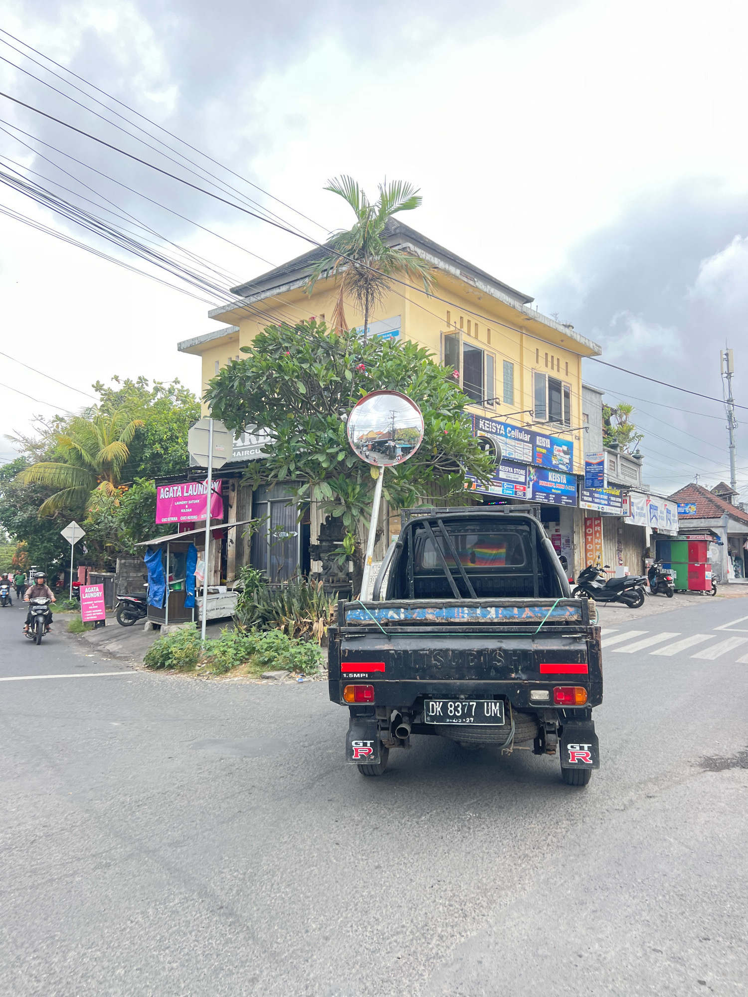 The image depicts a street scene in Denpasar, Indonesia, on an overcast day. In the foreground, the rear of a black pickup truck with a license plate reading "DK 8377 UM" is visible. The truck has a distinctive red "GT-R" emblem on its rear fender. To the left, a person is riding a motorcycle. Behind the truck, several businesses are visible, including "AGATA LAUNDRY" with pink signage and "KEISYA CELLULAR" with blue and white signage. Trees and foliage line the street, and utility wires crisscross the sky. A convex mirror is mounted on a pole, reflecting the street scene. Further down the street, more shops and vehicles, including other motorcycles and a red and green structure, are present. A cell tower can be seen in the background. The road is asphalt, and a crosswalk is painted on the surface.