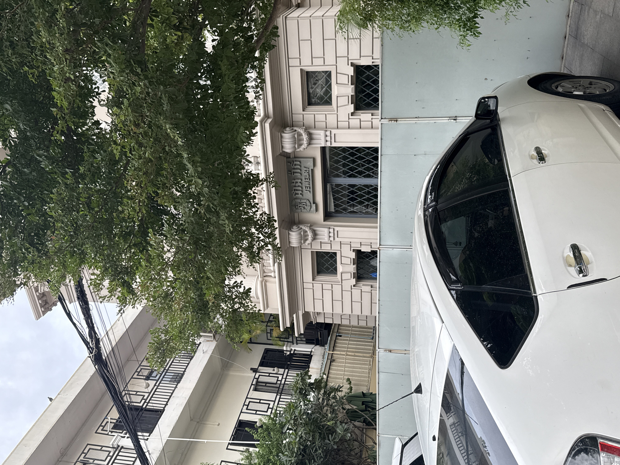 The image, captured in Khan Daun Penh, Cambodia, depicts a street scene dominated by a white car parked in the foreground. Behind the car, a light-colored building with classical architectural elements stands prominently. The building features decorative stone carvings around its entrance and windows, with a sign above a window displaying text in what appears to be Khmer script and the English word "JEWELRY". A large tree with green foliage partially obscures the upper portion of the building. To the left of the main building, a lighter colored structure with multiple balconies and dark railings is visible. Overhead, a complex network of electrical wires is strung across the frame. The overall environment suggests an urban setting, likely in a commercial or residential area. No people are visible in the image.