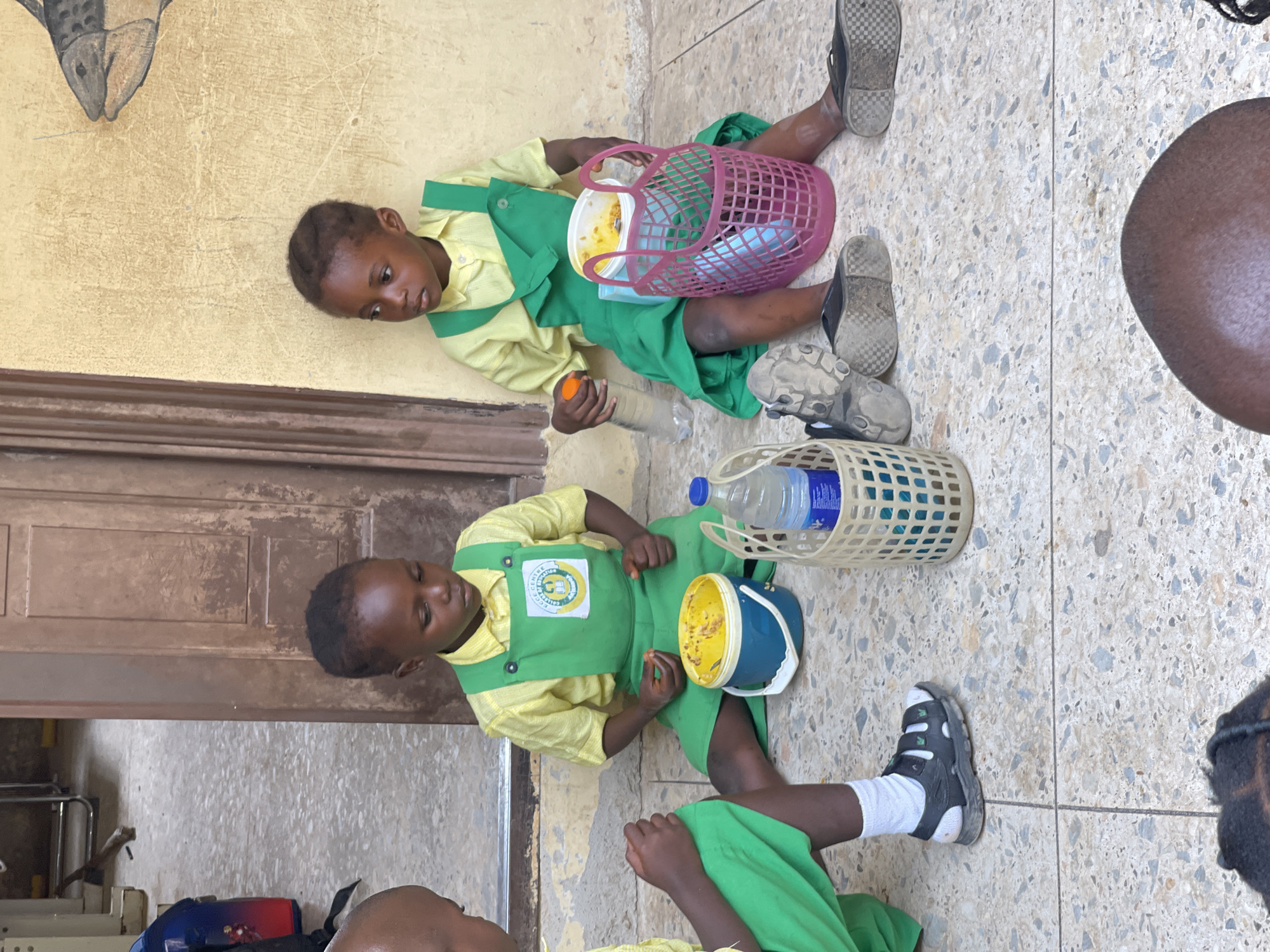 In Akwanga, Nigeria, two young children, appearing to be students, are seated on a terrazzo floor. The child on the left, wearing a yellow shirt and green pinafore, is looking down and holding a small object. Beside them is a blue bucket with a yellow lid containing food. The child on the right, also in a yellow shirt and green pinafore, is looking to the left and holding a clear plastic bottle with an orange cap. A pink woven basket, a beige woven basket, and another clear plastic bottle are also visible. The background features a textured yellow wall with a painted fish motif and a brown wooden door. The ground is a speckled terrazzo material, indicative of an outdoor or semi-outdoor school environment.