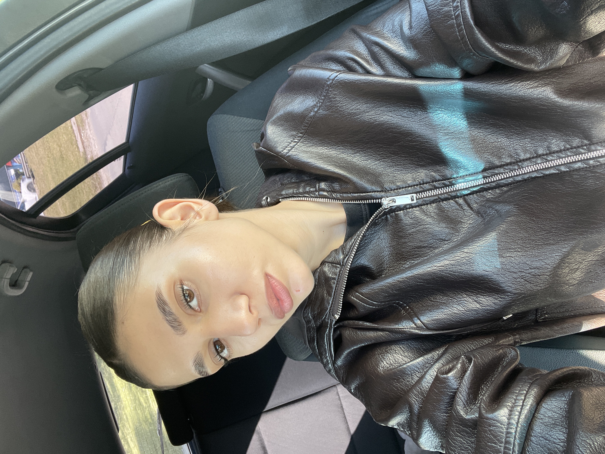 A young woman sits in the driver's seat of a car. She is wearing a dark brown leather jacket with a zipper that extends up to her neck. Her hair is pulled back. Light reflects off her skin. The car is parked and outside the window a grassy area is visible. There is no way to determine from this image if it is in Sofia, Bulgaria.