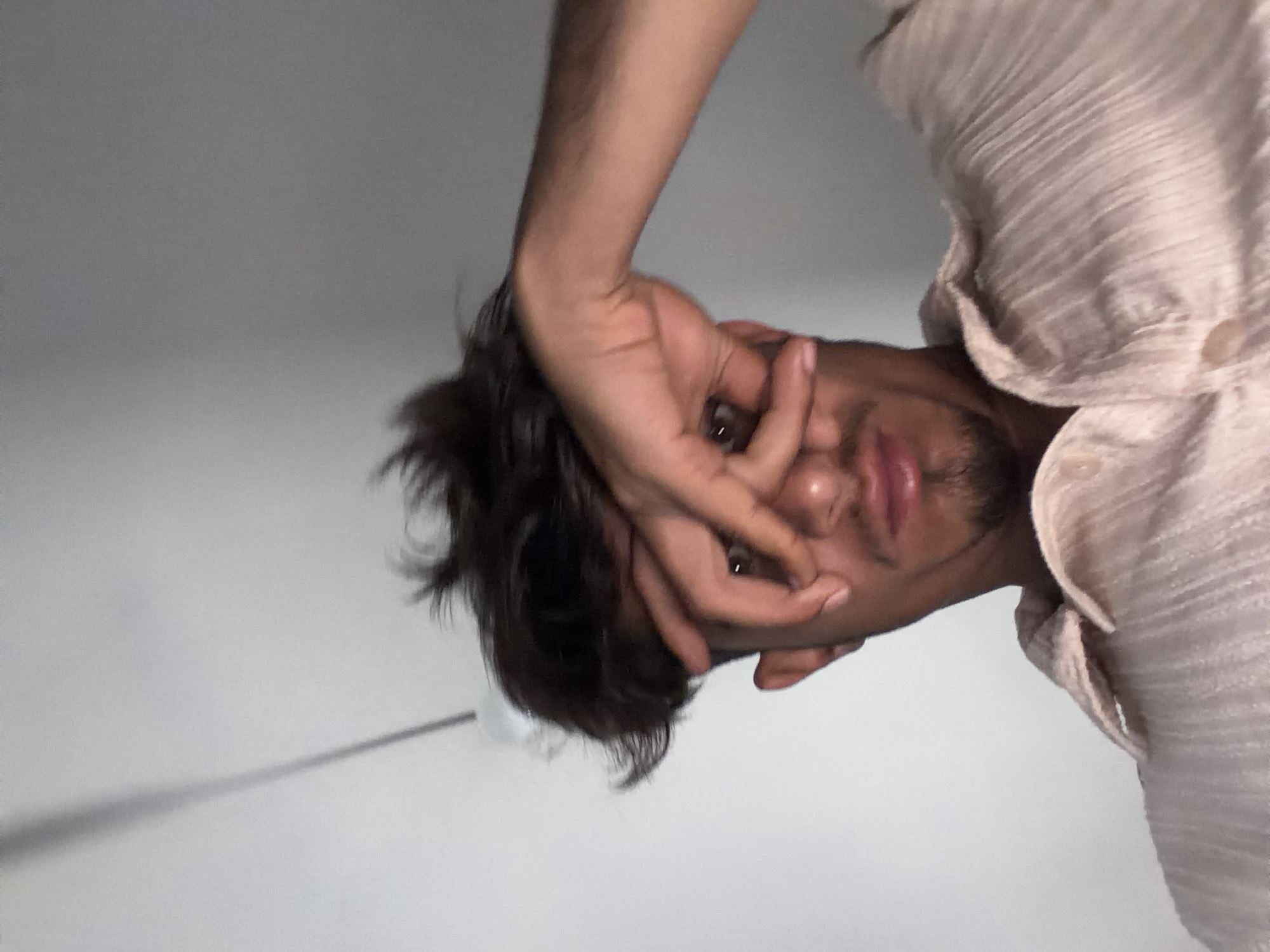 A person is viewed from an upside-down perspective. The person's face is partially obscured by their hand, with fingers covering one eye. The visible eye is dark brown. They have dark hair and a short beard. The person is wearing a light-colored, textured button-down shirt. The background appears to be a plain, light-colored wall. There is a faint, dark line extending from the person’s head to the left of the frame. There is no indication of the location being in Radnevo, Bulgaria.