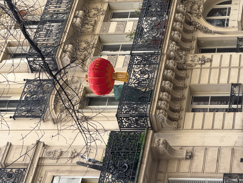 The image displays a multi-story Haussmannian-style building facade located in Paris, France. The building is constructed of light-colored stone, featuring several rows of windows and balconies with intricate architectural embellishments.
In the foreground, a large, spherical red Chinese lantern is suspended by a string from a bare, dark tree branch that stretches diagonally across the frame. The lantern is adorned with gold patterns and tassels, and legible Chinese characters, "法国华侨" (French Overseas Chinese), are printed in gold on its surface.
The building exhibits classic Parisian architectural elements including ornate black wrought-iron balcony railings with decorative scrolls and geometric patterns. These balconies are supported by multiple sculpted stone corbels, some featuring mascarons or other relief carvings. Windows are tall and narrow, some with white louvered shutters, while others show reflections of the surrounding environment. On one of the lower visible balconies, a dark green tarp or cover is present, alongside some sparse green foliage. No people are visible. The sky appears overcast, providing diffused light.