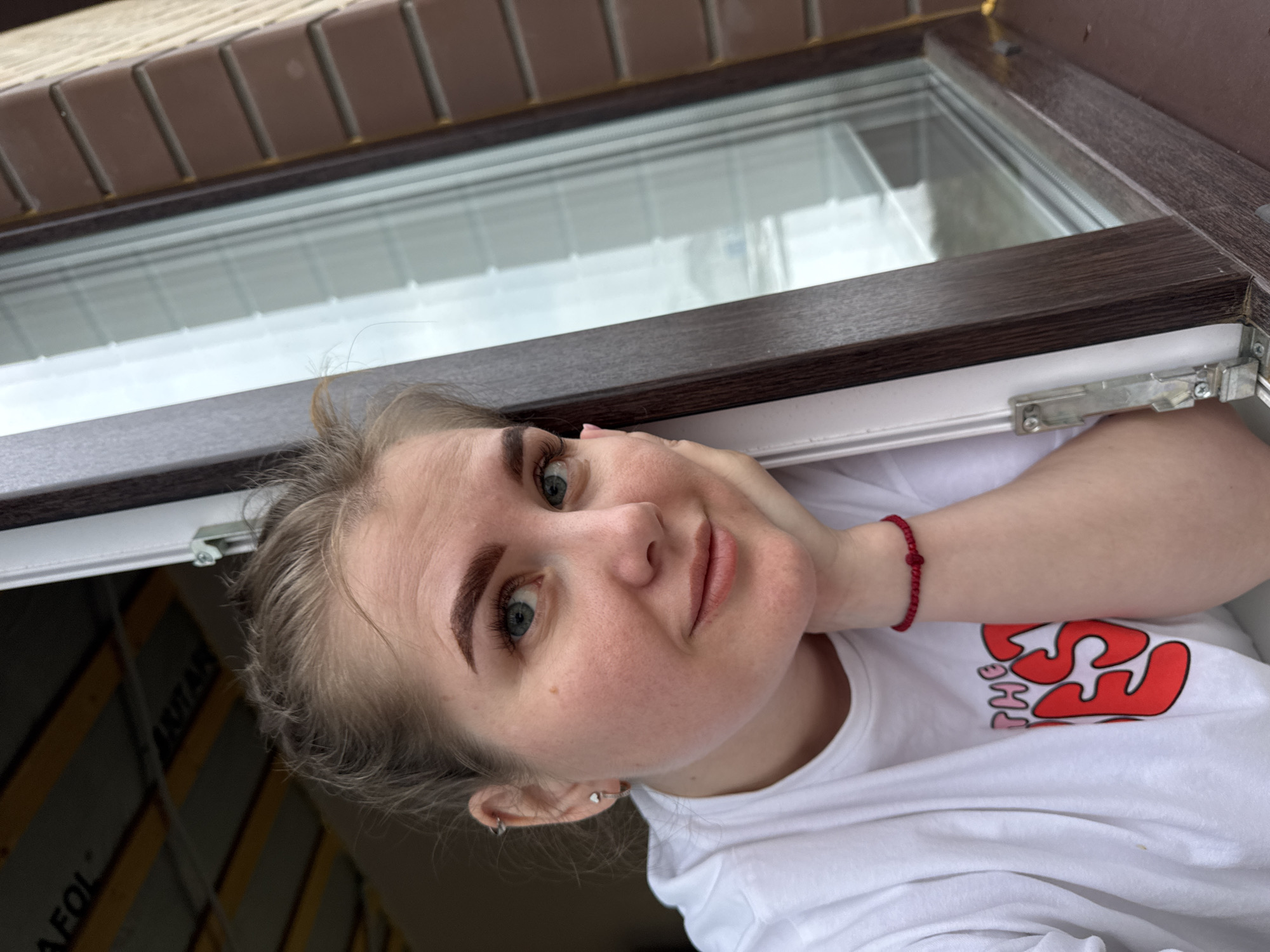 A young woman with fair skin and light brown hair is looking at the camera from below, resting her head on her hand. She has visible eyebrows and blue eyes. She wears a white t-shirt with a red design and a red beaded bracelet on her left wrist. She is positioned near a window frame with a brown wooden finish. The background through the window appears to be outdoors. The image is taken from a low angle, looking upwards. The window and frame are the primary surrounding elements. Based on the prompt, the location is Gornyi (Dvurechenskoe S/P), Russia.