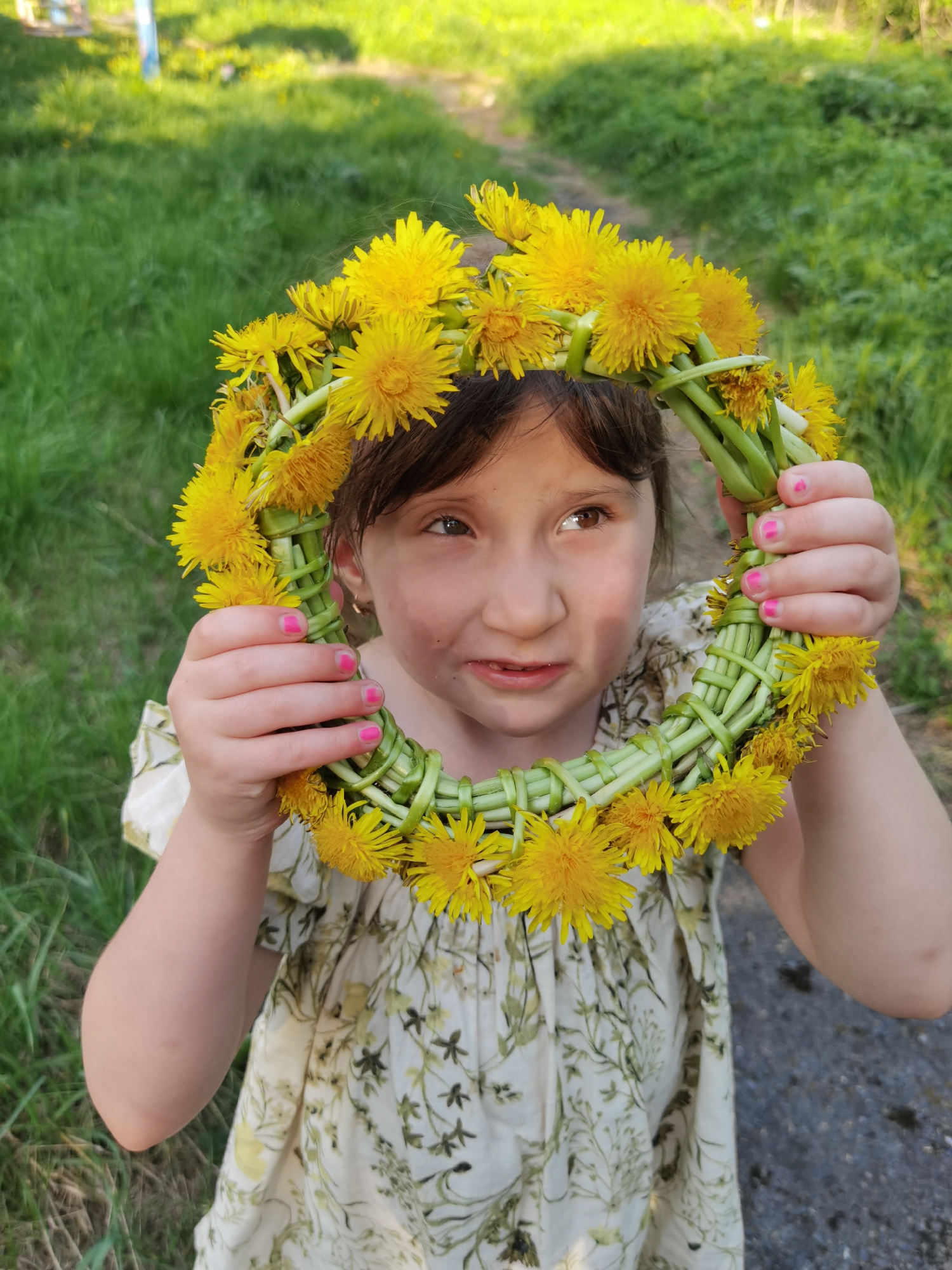 A young girl with brown hair and pink fingernails is holding a wreath made of dandelions in front of her face. She is wearing a light-colored dress with a floral pattern. The background consists of green grass and a gray paved area. The location is likely outdoors during daylight hours. The city of Perm, Russia is not evident. The main actions are the girl holding the wreath and looking off to the side.