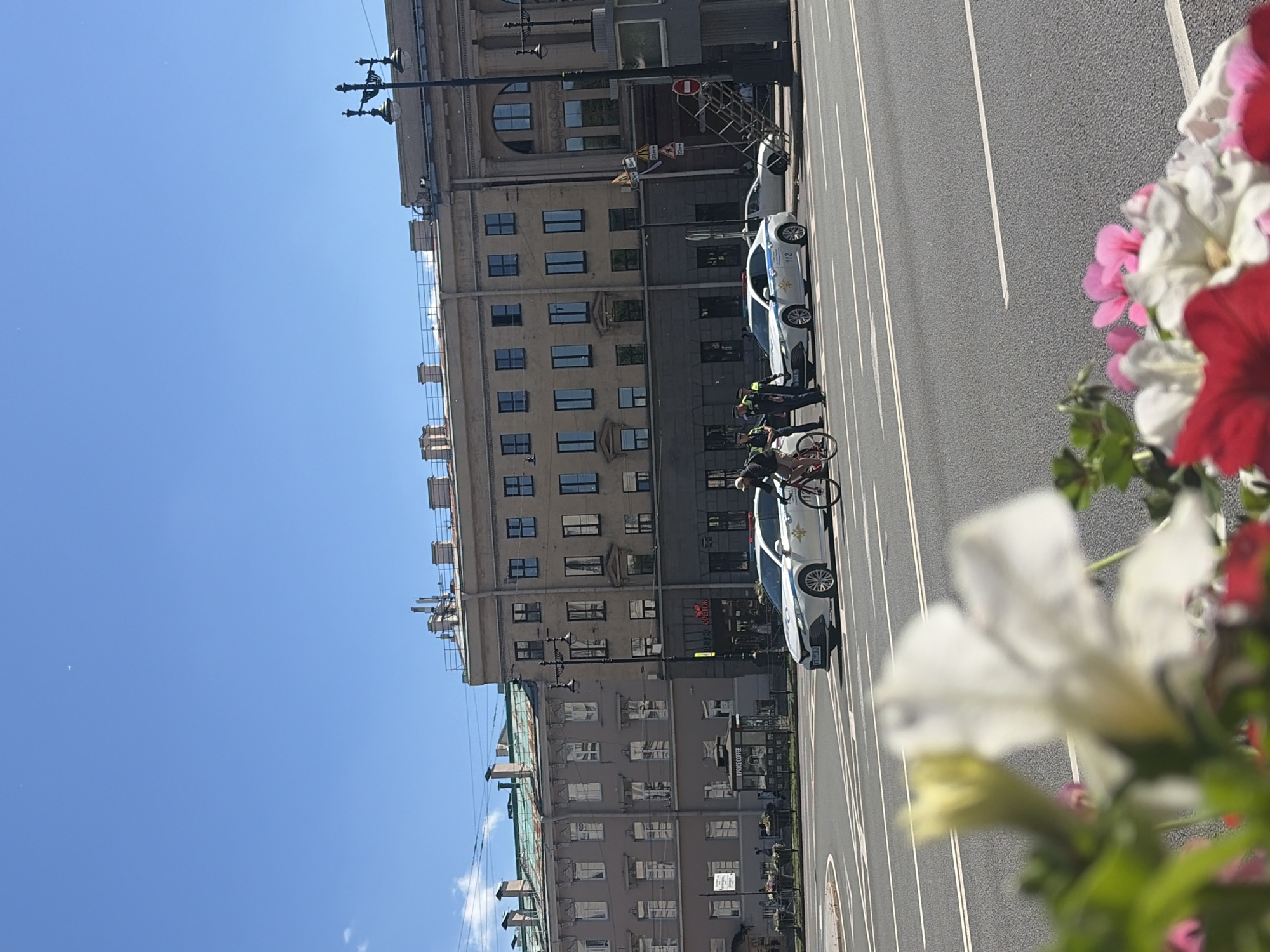The image depicts a street scene in Saint Petersburg, Russia, with the iconic architecture of the city in the background. The foreground is blurred by flowers, but the mid-ground reveals a street with multiple lanes of traffic. A white police car with flashing lights is present, and two police officers are interacting with a cyclist who is on a bicycle. Another white car is parked behind the police car. The sky is a clear blue with a few wispy clouds. The buildings in the background are multi-story structures with a classical architectural style, characteristic of Saint Petersburg. One building has a green roof, suggesting it might be an older structure. A lamppost with intricate detailing stands to the right of the frame. The overall impression is of a bright, sunny day in a major urban environment.