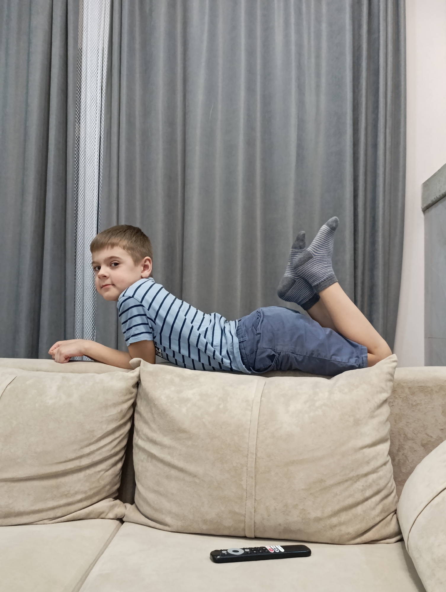 A male child, estimated to be between 6 and 10 years old, is depicted lying prone on a light beige upholstered sofa in an interior room in Chaikovsky, Russia. The child wears a light blue t-shirt with dark blue horizontal stripes, blue shorts, and grey socks with light stripes. His upper body is propped on his forearms, with his head turned slightly to his left, looking directly towards the camera with a neutral expression. His legs are bent at the knees, with his feet raised and crossed at the ankles. A black remote control with various colored buttons rests on the sofa surface in the foreground, positioned in the lower right quadrant of the image. The background consists of floor-to-ceiling light grey curtains, with a section of a white patterned sheer curtain visible to the left of the child. A light-colored wall section is visible in the upper right.