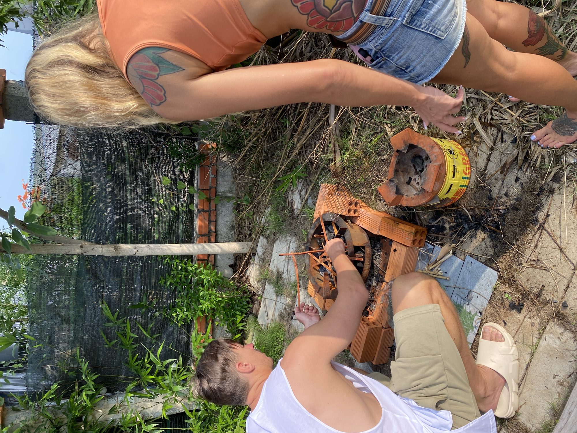 In Mũi Né, Vietnam, a man wearing a white tank top and light tan shorts kneels on the ground, tending to a small charcoal grill. He is actively arranging sticks and charcoal within the grill's metal chamber. Beside him, a stack of unglazed terracotta bricks is visible. A woman with blonde hair, wearing an orange crop top and denim shorts, stands to his right, her back to the camera. She has floral tattoos on her upper arm and thigh. The environment is outdoors, with dry grass, bamboo fencing, and a brick wall forming the backdrop. The ground is a mix of dirt, sand, and discarded plant matter. The sky is visible in the background, indicating daylight.