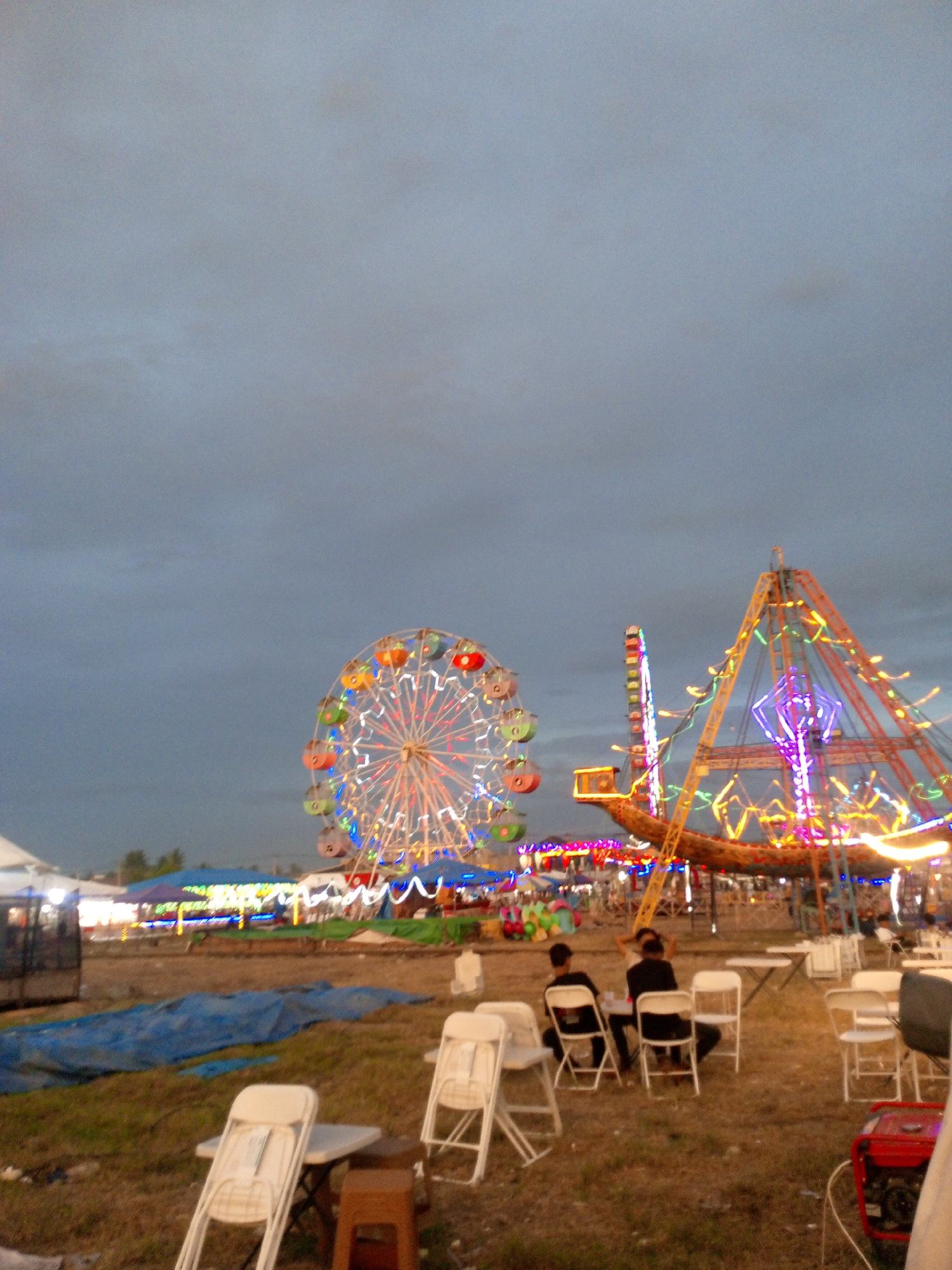 The image depicts an outdoor fairground scene in Blangcot Tunong, Indonesia, under a cloudy twilight sky. In the background, a brightly lit Ferris wheel and a triangular amusement ride with elaborate light decorations are prominently displayed. Various stalls and tents are visible behind these rides, illuminated with colorful lights. In the foreground, several white folding chairs are arranged around small tables on a grassy, uneven ground. Two individuals are seated at one of the tables, facing away from the camera, engrossed in conversation. A red portable generator is positioned in the lower right corner of the frame. The overall atmosphere suggests a festive event taking place as evening descends.