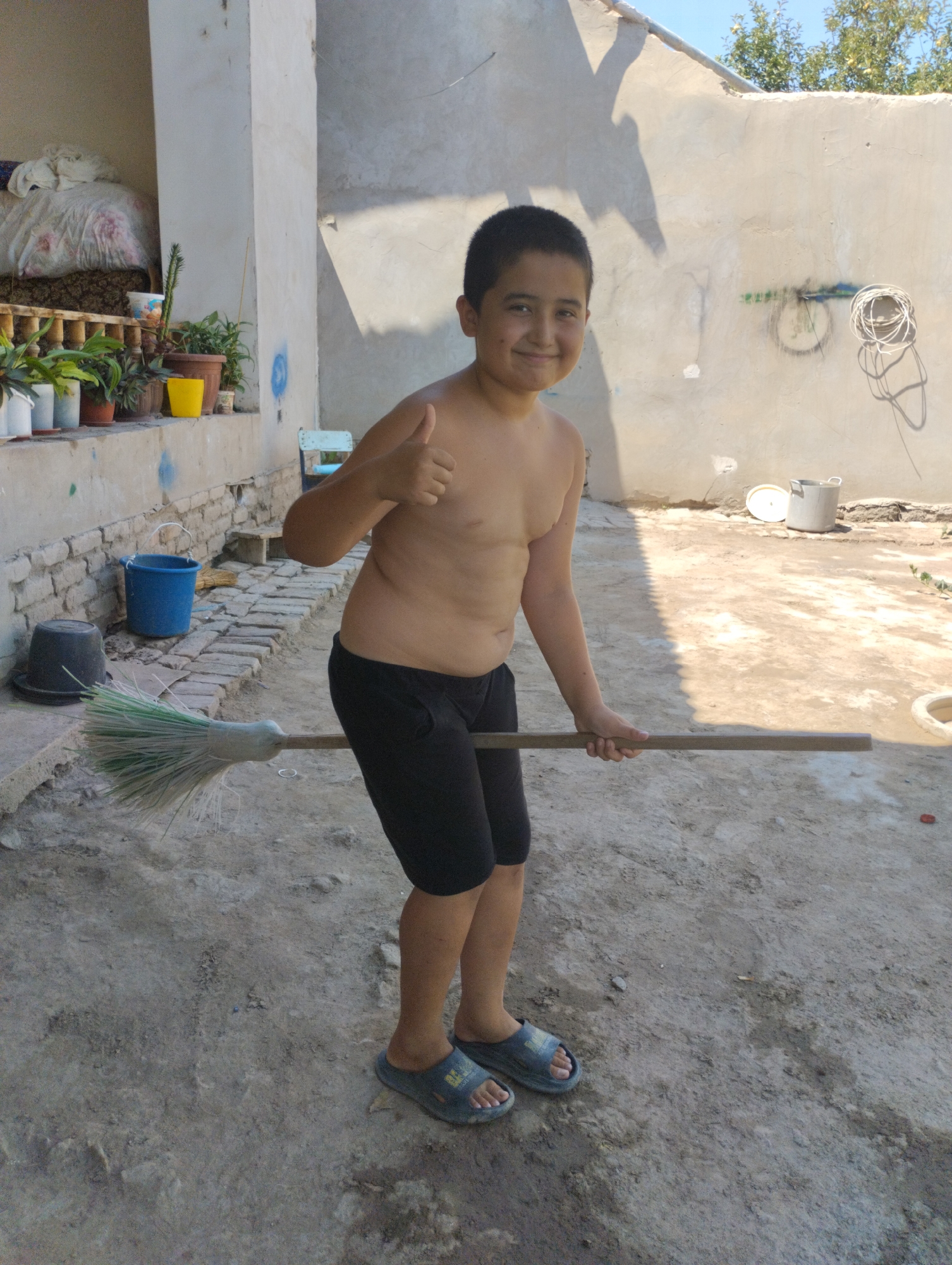 A young boy, appearing to be of Central Asian descent, stands in a sunlit outdoor courtyard in Yangibazar, Uzbekistan. He is shirtless, wearing black shorts and blue sandals. The boy is holding a broom horizontally with both hands and giving a thumbs-up with his right hand, smiling broadly at the camera. The broom has a light-colored wooden handle and a green and white bristled head.
The courtyard has a dirt and gravel ground. To the left of the boy, a low wall with bricks and a concrete top runs along a building. On this wall, several potted plants are visible, including a bright yellow pot and a white pot with red flowers. Behind the plants, a white sheet is draped over something that resembles a bed. Next to the wall, a blue bucket and a black bucket with a lid are placed on the ground, with a broom leaning against the wall near the blue bucket.
The background features a plain, light-colored wall with some graffiti and a coiled white rope hanging from it. The wall casts a long shadow of the boy onto the ground, indicating it is likely mid-afternoon. The sky is clear and blue, suggesting fair weather. In the distance, the top of some trees can be seen. The overall scene depicts a casual, everyday moment in a rural or semi-rural setting. No visible text is present in the image.