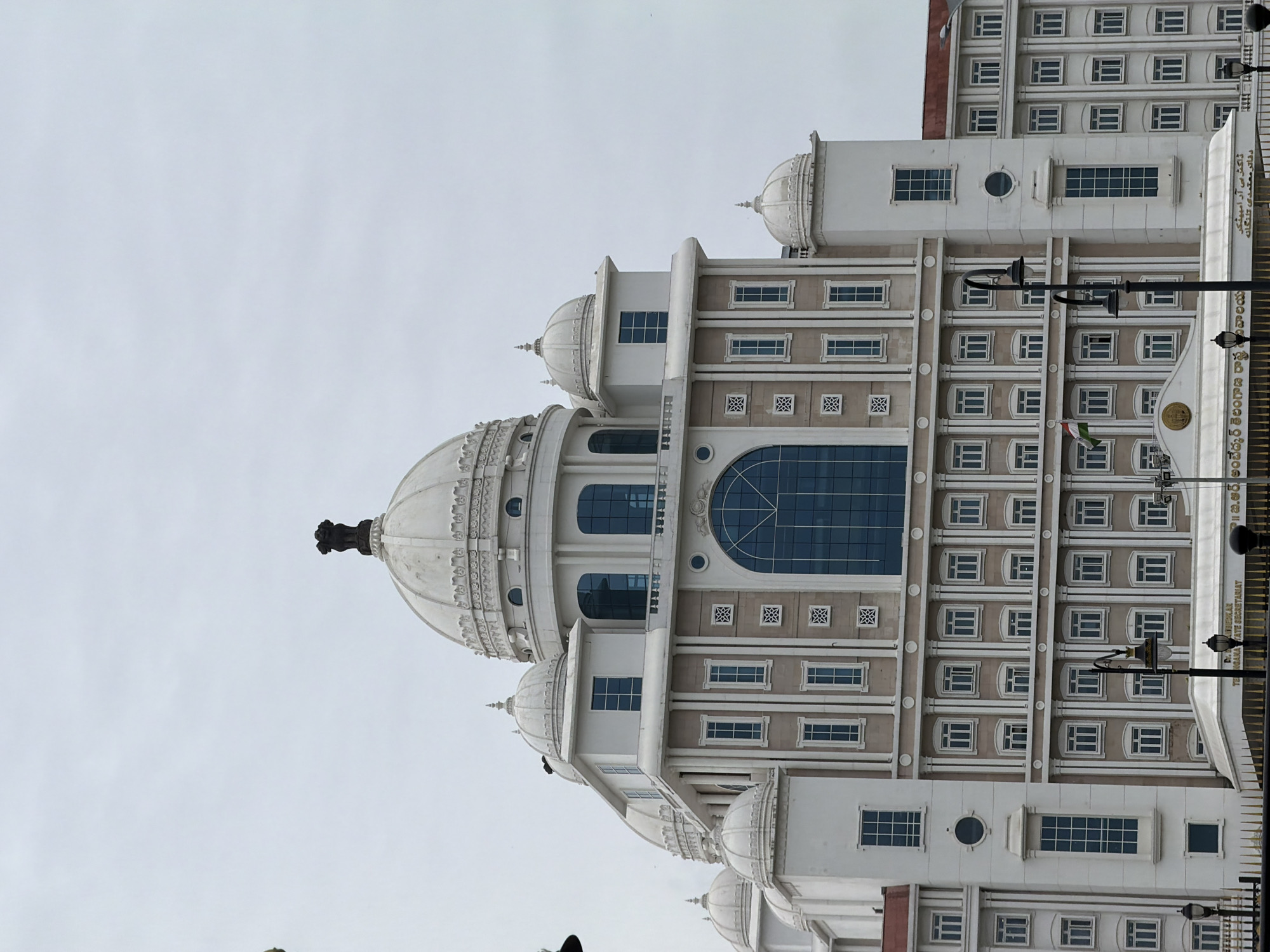The image captures the Telangana Secretariat in Hyderabad, India, under a cloudy sky. The imposing structure features multiple domes, a prominent central dome topped with the Ashoka Pillar, and a facade with numerous windows. The building's design incorporates classical architectural elements with a modern aesthetic. In the foreground, ornate lampposts and a golden railing suggest a formal setting. The presence of the Indian flag adds a patriotic touch to the scene. The overall impression is one of grandeur and officialdom.