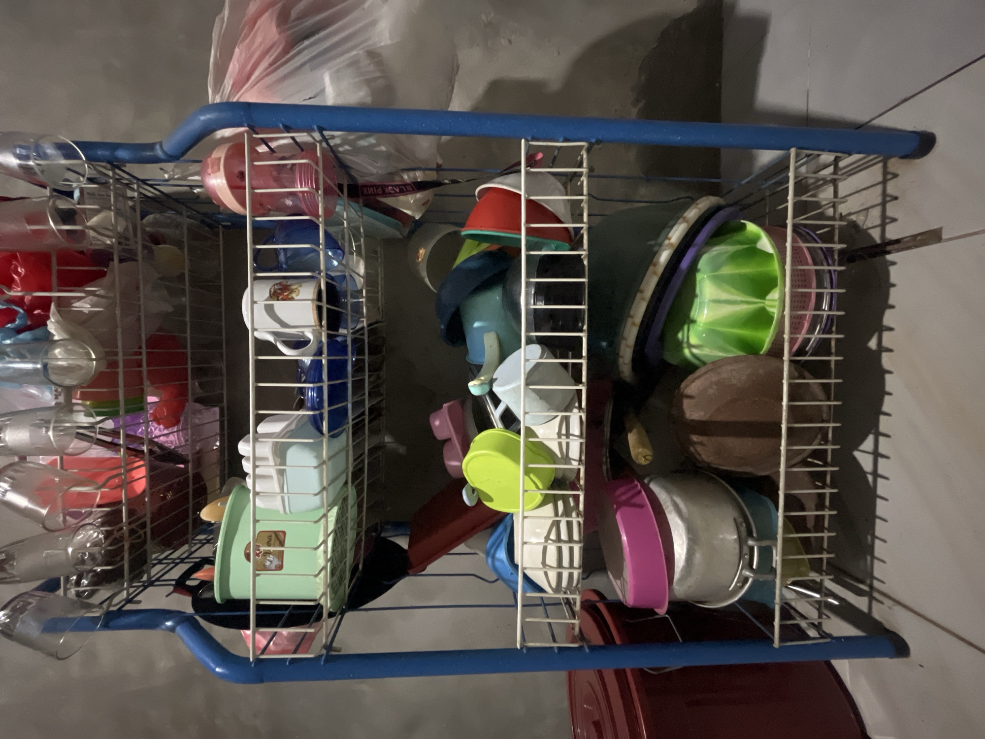 An indoor, medium shot captures a blue metal dish drying rack filled with various kitchenware. The rack is positioned against a light-colored, textured wall, with a tiled floor visible below. On the top shelf, clear drinking glasses and some red and pink plastic items are stacked. The second shelf holds green and blue plastic containers, white cups, and a pink baby bottle. Below that, more plastic containers, including a bright green one, are visible, along with a dark blue object and some red and white bowls. The bottom shelf contains large silver pots with colorful lids, a brown round object that appears to be a coconut, and green and purple bowls. A red plastic container is partially visible to the left of the dish rack. The lighting suggests it is nighttime or the room is dimly lit. This scene is likely located in Kebumen, Indonesia, as indicated by external context, suggesting a typical kitchen or utility area in a residential setting.