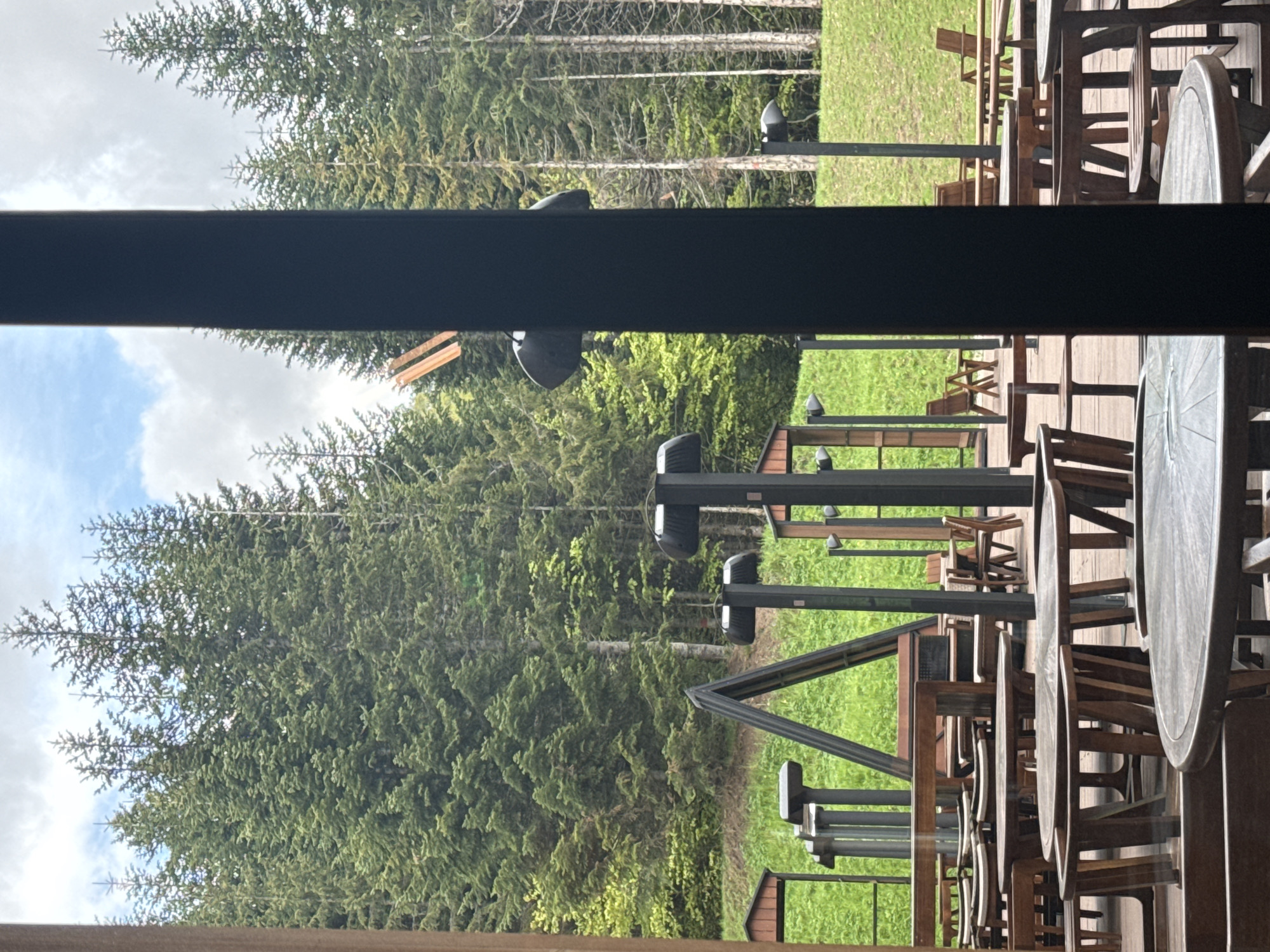 The scene is outdoors, likely a patio or deck setting, framed by a dark horizontal beam suggesting a partial view from inside a building. Several wooden tables and chairs are arranged on a wooden deck, suggesting a dining or gathering area. In the background, beyond the deck, is a dense green forest with tall trees reaching towards a sky with some clouds. There are also some lampposts visible between the trees. The overall impression is of a restaurant or lodge with outdoor seating, bordered by forest. There is no visual information that identifies the specific location as the city of Katun Bačko brdo, Montenegro.