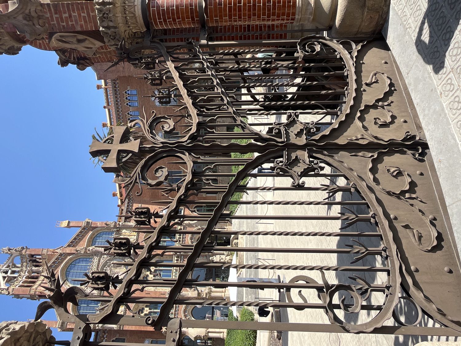 The image, if rotated correctly, captures an ornate wrought-iron gate in the foreground, providing a view of a grand architectural building behind it under a clear blue sky.
The gate is highly detailed, featuring elaborate scrollwork, twisted iron bars, and embossed organic patterns. A prominent Maltese cross design adorns the upper right section of the gate, while smaller, decorative elements resembling stylized insects or gargoyles are affixed along its horizontal railings.
Behind the intricate gate stands a magnificent brick building, characteristic of Modernista architecture. It showcases multiple levels, large arched windows with decorative frames, and rich sculptural or relief details along its cornices and facades. Green foliage, including palm fronds, is visible peeking through the gate and framing parts of the building, suggesting well-maintained landscaping.
The ground beyond the gate is paved with light-colored tiles, some displaying a repeating pattern reminiscent of a four-leaf clover or stylized flower. A clear, distinct shadow, likely of a person's head and arm holding a device, is cast onto these patterned tiles in the lower right portion of the frame. The bright, clear blue sky and strong shadows indicate a sunny day, likely midday or early afternoon. The scene evokes a sense of historical grandeur and architectural beauty, consistent with landmarks found in Barcelona, Spain. No visible text is present in the image.