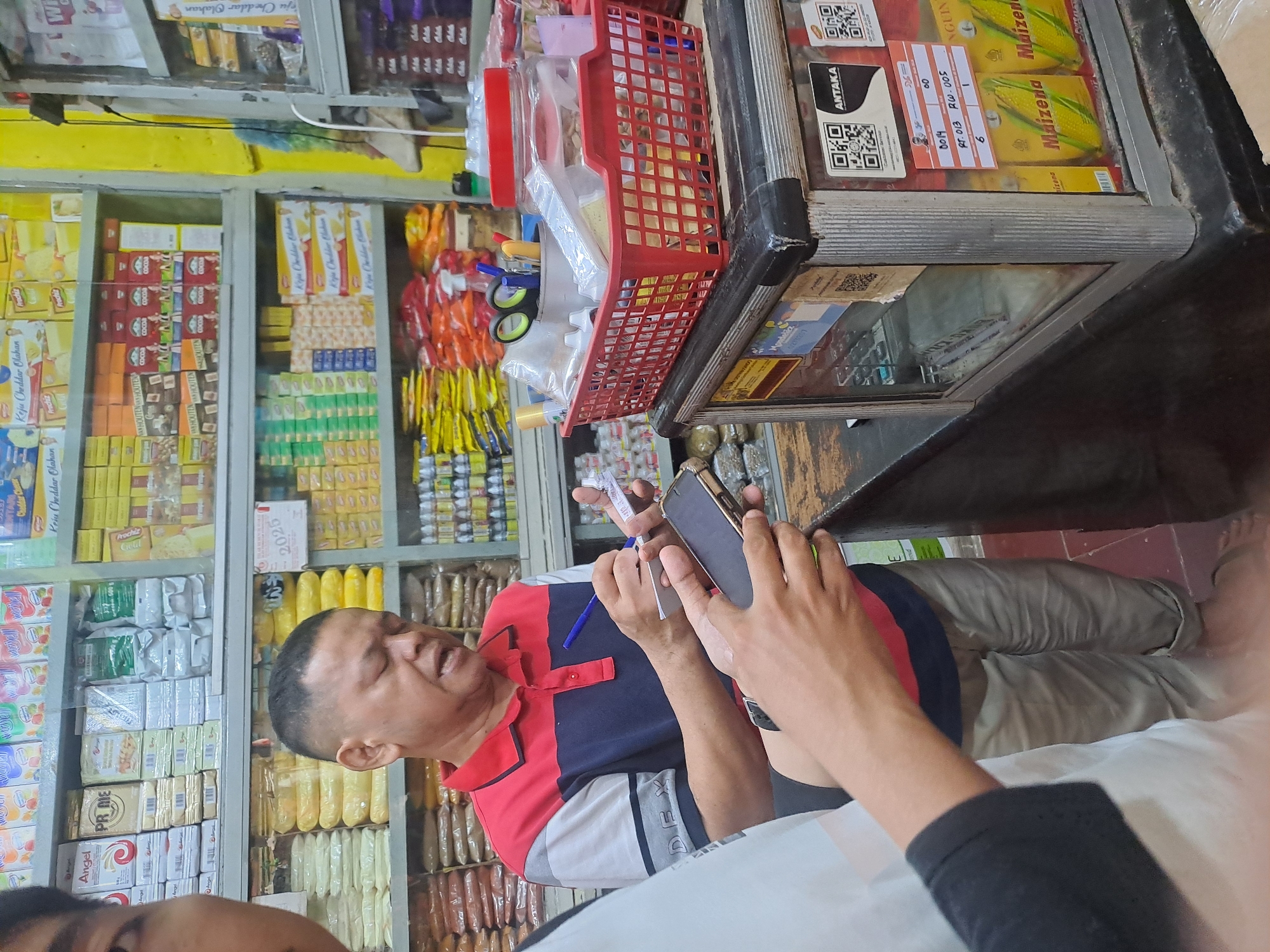 The image depicts an indoor scene, likely a small retail store or warung, in Palembang, Indonesia. In the foreground, a man wearing a red, blue, and gray collared shirt is holding a blue pen and writing on a small piece of paper. He is looking down at his task. To his left, another person's hands are visible, holding a smartphone and a piece of paper, suggesting an interaction between the two individuals, possibly related to a transaction or inventory.
Behind the man, shelves are stocked with various packaged goods. These include items in yellow and red packaging, with some visible text indicating brands or product types. The store appears to be modestly equipped, with simple shelving units and a counter area. A red plastic basket containing office supplies like scissors, pens, and bottles is placed on top of a display case. The display case itself has a glass front, revealing items inside, and features various labels and QR codes on its exterior. One label is a price list or order form with handwritten entries. The overall ambiance suggests a local, everyday commercial setting.