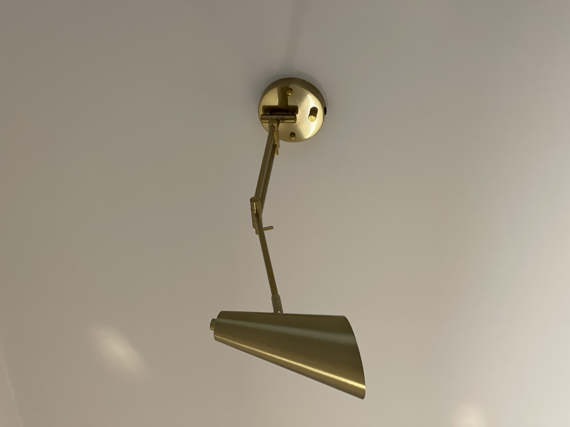 A low-angle, medium shot captures a gold-colored metal desk lamp mounted on a white ceiling. The lamp features a circular base attached to the ceiling and an adjustable arm extending downwards. The lampshade is conical in shape, tapering towards the bottom, with a reflective surface. The ceiling appears smooth and matte, reflecting light in various areas. No people or specific location details like Izhevsk, Russia are identifiable in the image.
