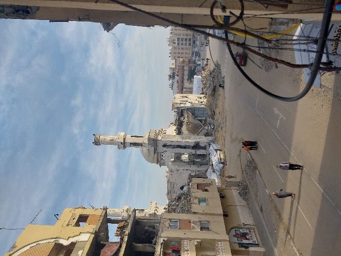 An elevated street view depicts a scene of extensive urban destruction in Khan Yunis, Palestinian Territory, under a partly cloudy sky. In the mid-ground, the Great Mosque of Khan Yunis appears severely damaged; its dome is partially intact, and the minaret stands but shows damage to its upper sections, while large portions of the surrounding mosque structure have collapsed into rubble. To the left, a multi-story building exhibits significant structural damage, with walls missing, interiors exposed, and debris scattered; laundry is visible hanging on a line from a balcony on one of its upper floors. On the far right foreground, a damaged building facade with exposed wires and structural elements partially frames the image.
The asphalt street in the foreground, marked with white lines, is mostly clear of heavy debris. Two individuals walk away from the camera in the immediate foreground; one wears a light-colored thobe or robe, and the other wears dark clothing. Further down the street, a group of approximately five individuals stands together near the center. Additional figures are visible walking among the rubble piles and structures in the background. Extensive piles of concrete and building debris are present throughout the scene, particularly around the damaged mosque and adjacent structures. A small red vehicle is parked on the right side of the street in the mid-ground. Other multi-story buildings, some appearing less damaged, are visible in the distant background.