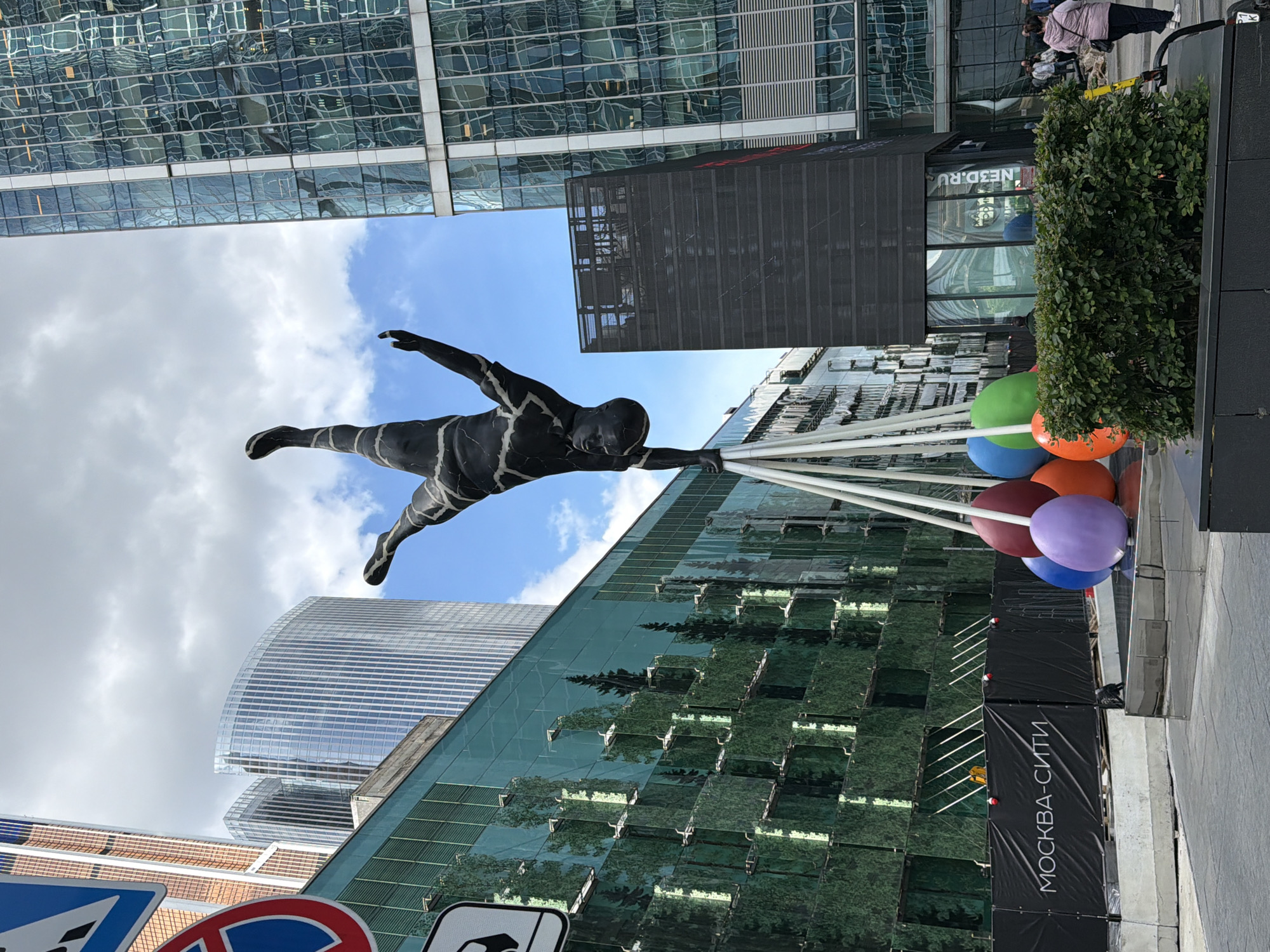 The image depicts a statue of a person flying while holding onto several balloons in Moscow, Russia. The statue is dark and has an abstract, striped pattern. The balloons are multicolored including green, blue, orange, red, and purple. The background features modern skyscrapers, some with reflective facades that mirror the sky and trees. The sky is partly cloudy. There are a few people near the top-right corner. A sign on a dark fence-like structure at the bottom reads "МОСКВА-СИТИ".