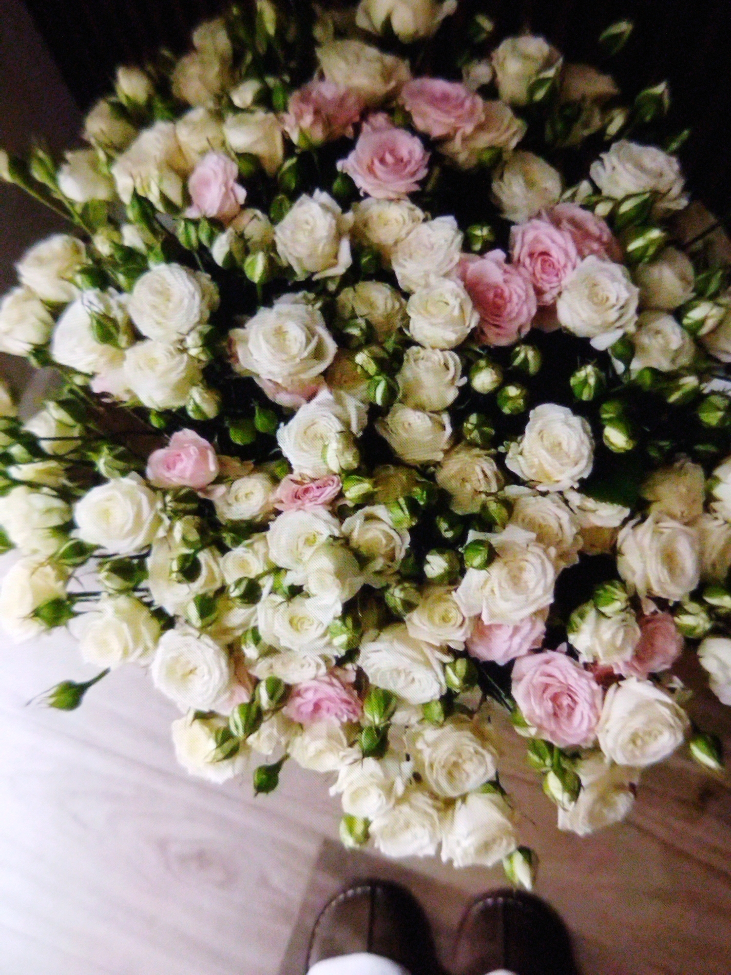 The image shows a close-up, overhead view of a large bouquet of cream and light pink roses with green buds. The roses are tightly packed and appear to be of a smaller varietal, possibly spray roses. The bouquet is resting on a light-colored wooden surface, possibly a floor or a table, which has visible grain and a matte finish. Some dark, out-of-focus shapes, which appear to be shoes, are visible at the bottom of the frame, suggesting a person is present. The lighting is dim, with some areas of the bouquet being brightly lit, creating a slight contrast between the roses and their shadows. The background is very dark and out of focus. The overall impression is one of a still life arrangement, possibly indoors. There is no visible text in the image.