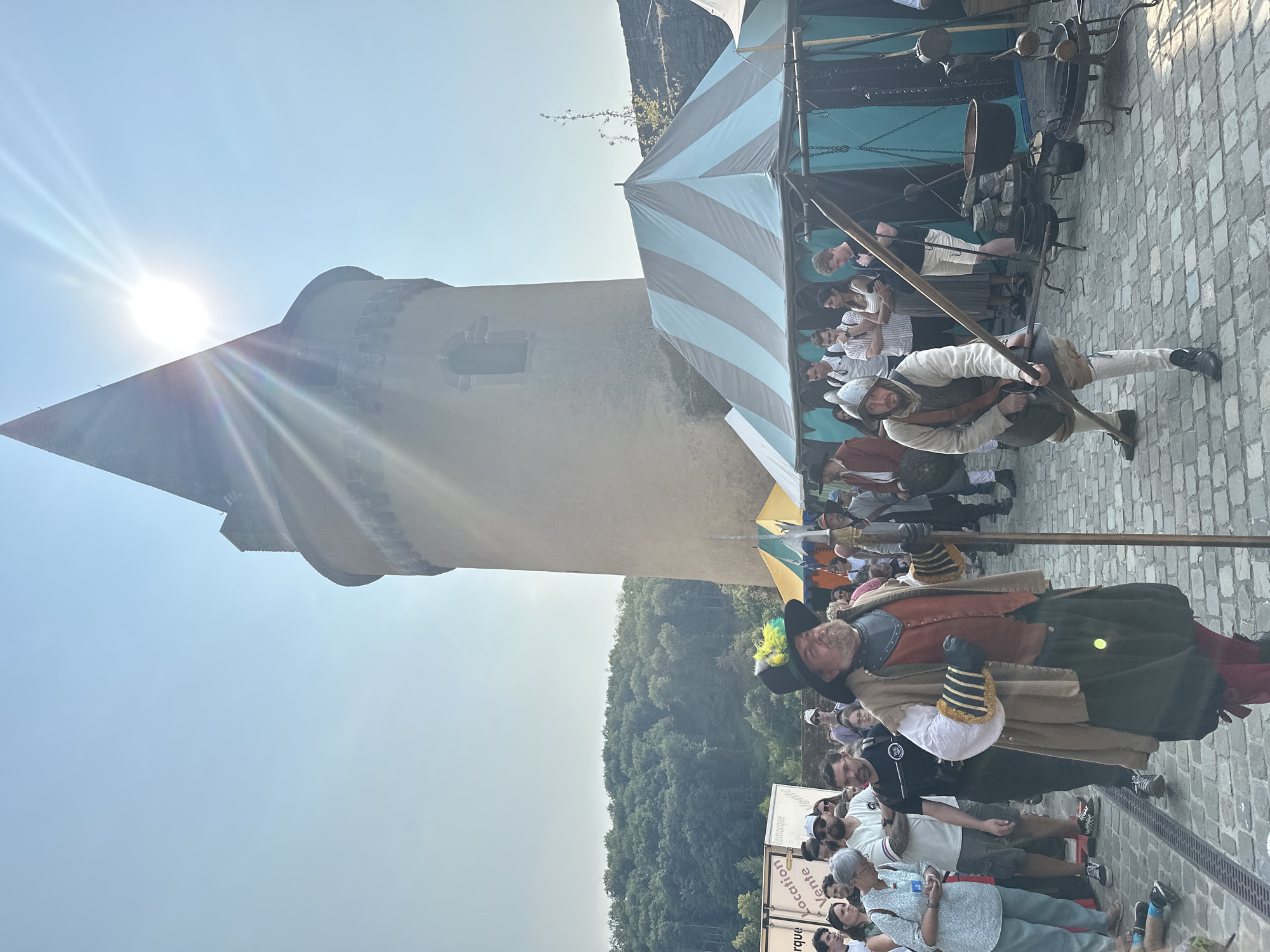 This image depicts a sunny outdoor scene at a historical reenactment event, likely held at or near the Vianden Castle in Vianden, Luxembourg. In the foreground, two men dressed in historical costumes are prominent. The man on the left wears a black hat adorned with feathers, a tan cloak over a red vest, and a green skirt-like garment. He holds a tall staff. To his right, another man in historical attire, wearing a helmet, a light-colored tunic, and knee-high stockings, holds a spear.
Behind them, a crowd of people in modern clothing are visible, suggesting they are spectators. Several tents with striped awnings are set up, one predominantly light blue and gray. In the background, a tall, cylindrical stone tower with a conical roof dominates the skyline. This structure, likely part of the Vianden Castle complex, is partially obscured by the bright sun which casts lens flares across the image. The ground is paved with cobblestones. The environment also includes a forested hillside in the far background and a clear blue sky with strong sunlight. Various historical-looking cooking vessels and equipment are displayed outside one of the tents. A trailer with French text that reads "Remorque Location Vente" is also visible on the left side of the frame.