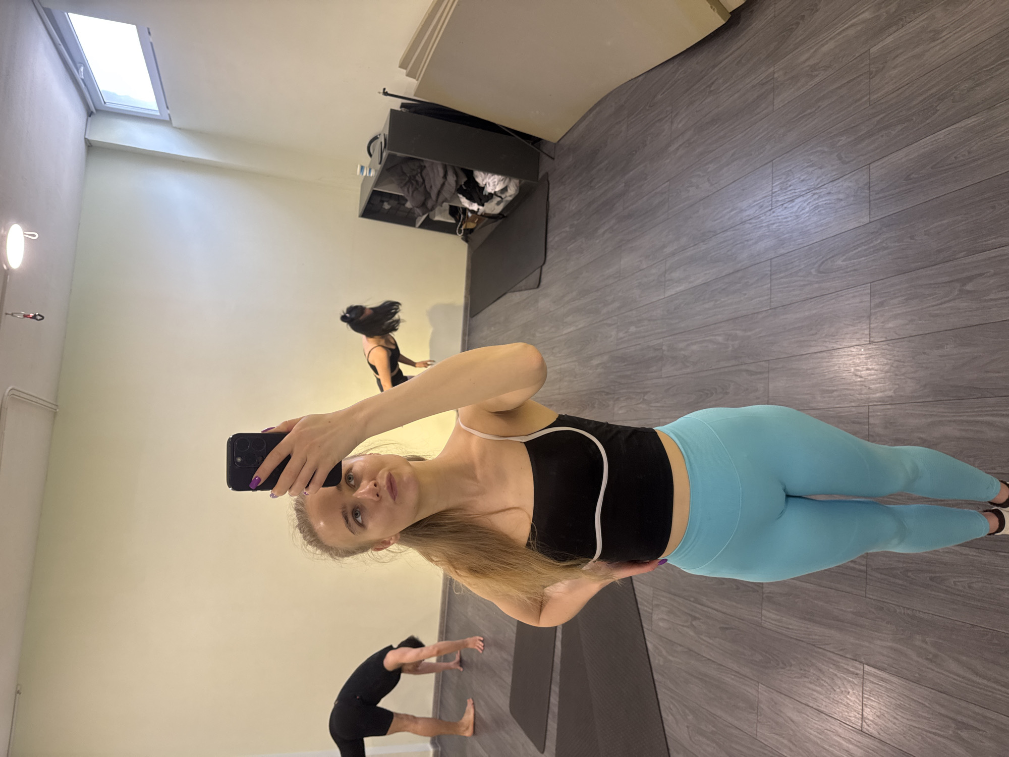 A young woman is captured in a mirror selfie in what appears to be a yoga or fitness studio. She is the central figure, holding a black smartphone with her right hand, showcasing purple nail polish. She has long, light blonde hair and a neutral expression, looking directly into the camera. She wears a black crop top with white trim and form-fitting light blue leggings.
In the background, two other individuals are visible. To the left, a person with dark hair, possibly a woman, is seen from the back, wearing a black top and dark leggings, seemingly in an exercise pose. Further back and to the right, another person, also in dark exercise attire, is bending forward on a dark yoga mat.
The room features light-colored walls and a grey wooden plank floor. A rectangular skylight or window is visible on the ceiling, providing natural light. A black shelving unit, appearing somewhat cluttered with items, is positioned against a back wall. Two dark yoga mats are laid out on the floor. A circular wall-mounted light fixture is visible on the left wall. The overall scene suggests an indoor fitness environment where multiple people are engaged in physical activity. There is no visible text in the image.