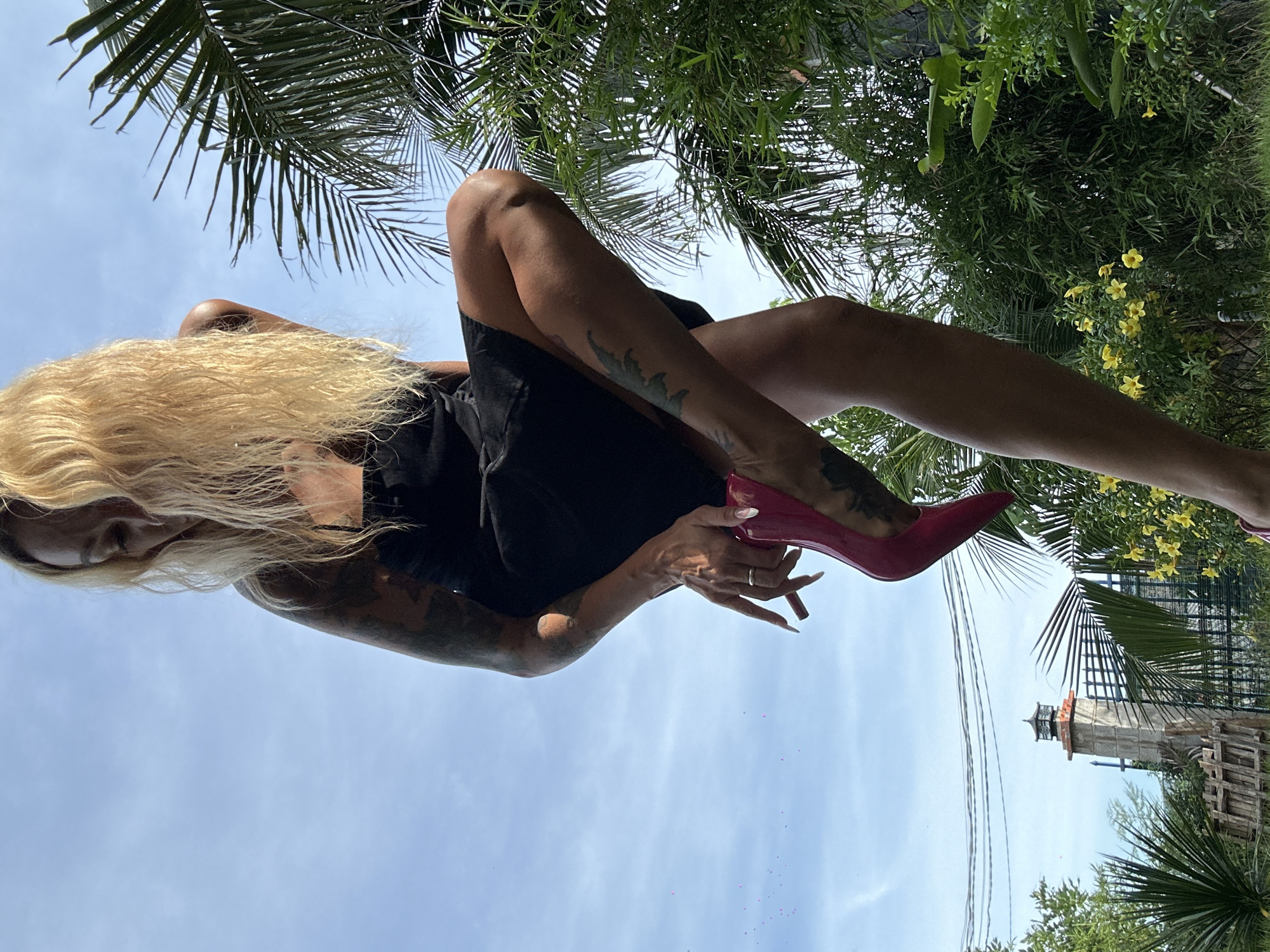 A woman with blonde hair and tattoos on her arms and legs is wearing a black dress and red high heels. She is outdoors in what appears to be a garden or lush green area with palm trees. In the background, there is a lighthouse-like structure, a fence, and power lines, suggesting an urban or semi-urban setting in Mũi Né, Vietnam. The sky is blue with some clouds. The woman is in the process of putting on or adjusting one of her shoes.
