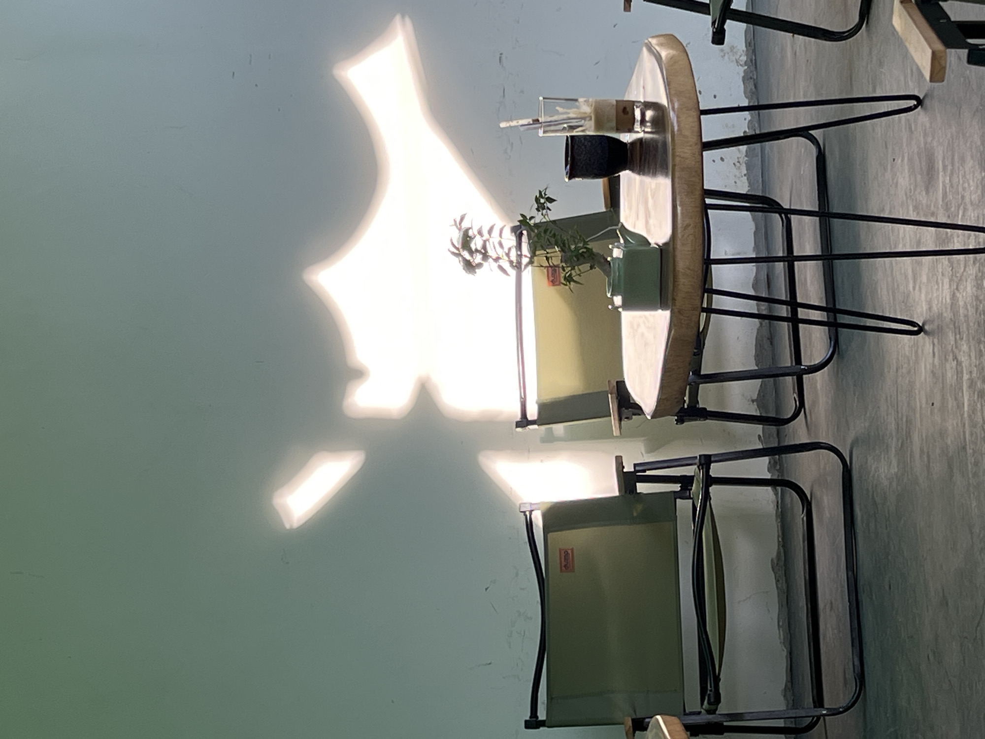 The image depicts an outdoor seating area, likely a cafe or restaurant, in Nha Trang Ward, Vietnam. The scene is set against a light blue wall marked with faint shadows. Two olive green camping-style chairs with black metal frames are visible. In the foreground, a small, irregularly shaped wooden table with thin black metal hairpin legs holds a small, light green teapot, a dark blue cup, and a tall glass with a straw containing a layered beverage. A small plant with green and red leaves sits between the teapot and the cup. The floor is a rough, grey concrete surface. Sunlight casts dramatic, abstract shapes on the wall behind the chairs, suggesting a window or a light source is filtering through an object. The overall impression is one of a quiet, relaxed setting.
