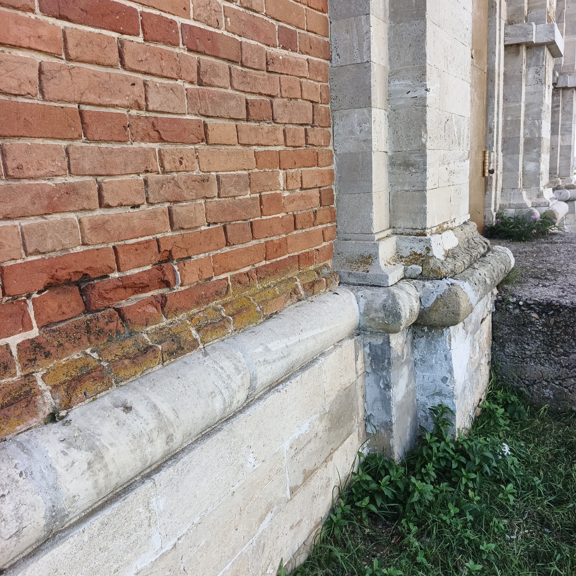 The image shows a detail of the exterior of a building, likely a historical one, with a brick wall on the left and stone architectural elements on the right. The brick wall is constructed of reddish-brown bricks, with some areas showing signs of wear and discoloration, including patches of yellow and black growth, possibly moss or lichen. A prominent white, curved stone element runs horizontally along the base of the brick wall. To the right, a large stone column or pilaster is visible, composed of stacked rectangular stone blocks. This stone structure has a decorative molding at its base, also made of stone, which is worn and shows signs of chipping. The overall impression is one of age and weathering. In the foreground and to the right of the stone structure, there is green grass with some small leafy plants and a few small white flowers. The location is stated to be in the city of Tugustomir, Russia.