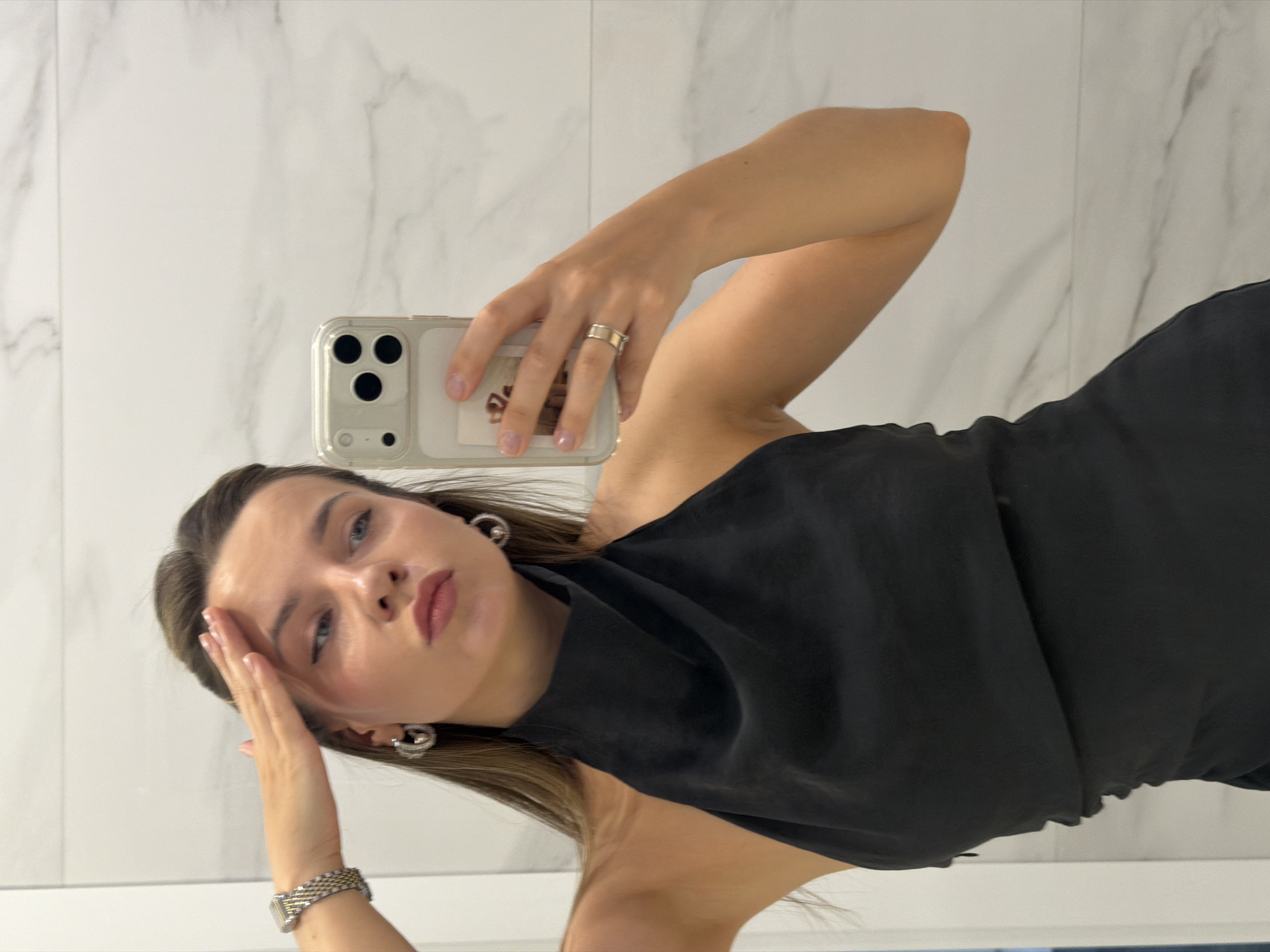 A young woman with long, brown hair, wearing a black halter top and silver hoop earrings, takes a selfie in a mirror. Her right hand is raised to her forehead, and she is wearing a watch on her left wrist and a ring on her right ring finger. She is holding a light-colored smartphone, which is also visible in the mirror. The background appears to be a bathroom with white marble tiles.