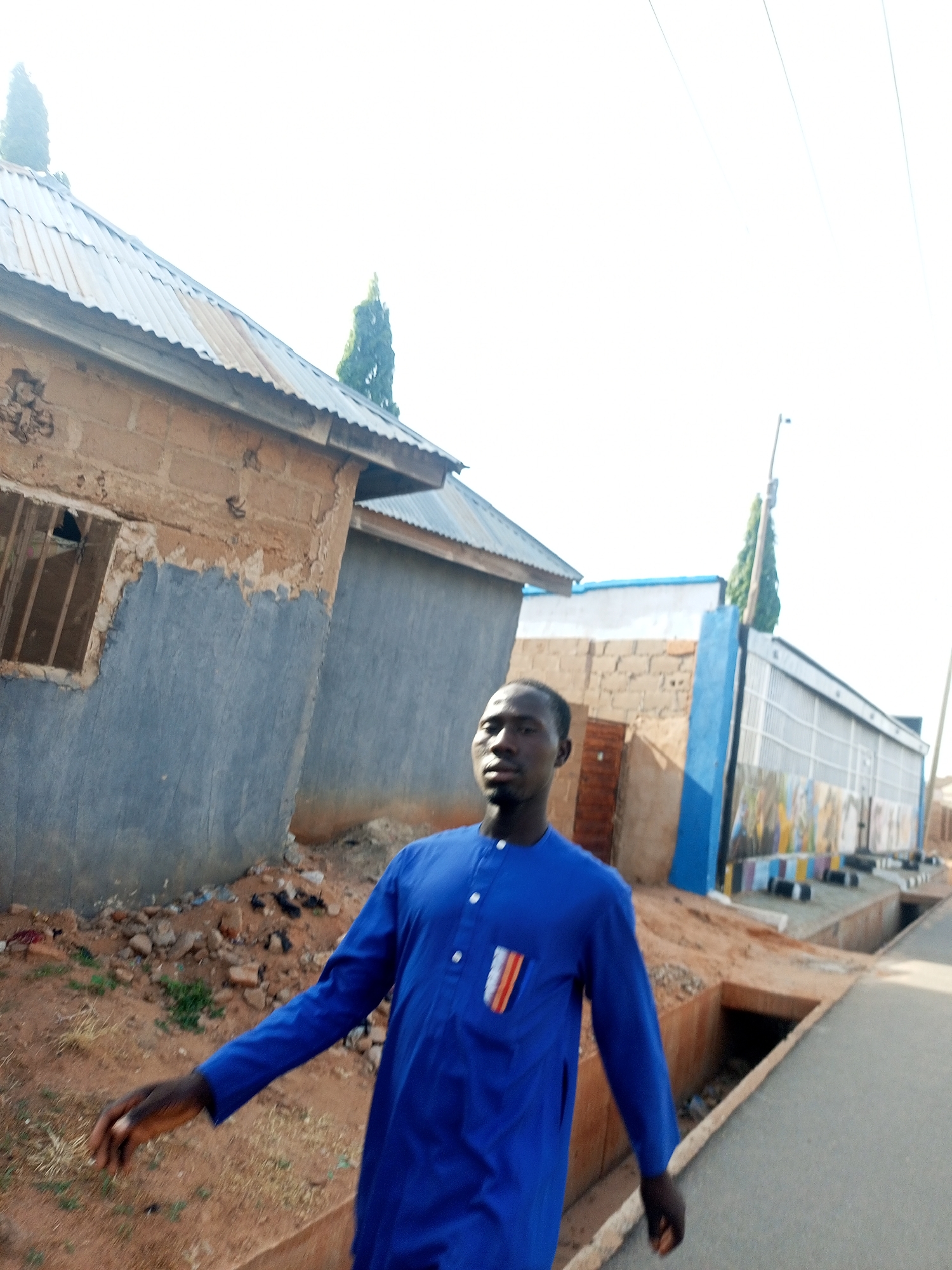 A man stands in the foreground, wearing a blue, long-sleeved shirt with a pocket square featuring red, white, and orange stripes. He is positioned on a sidewalk next to a gutter. Behind him are buildings of varying construction styles, including one with a corrugated metal roof and another with a painted blue facade. The environment appears to be a street in Tempure, Nigeria.
