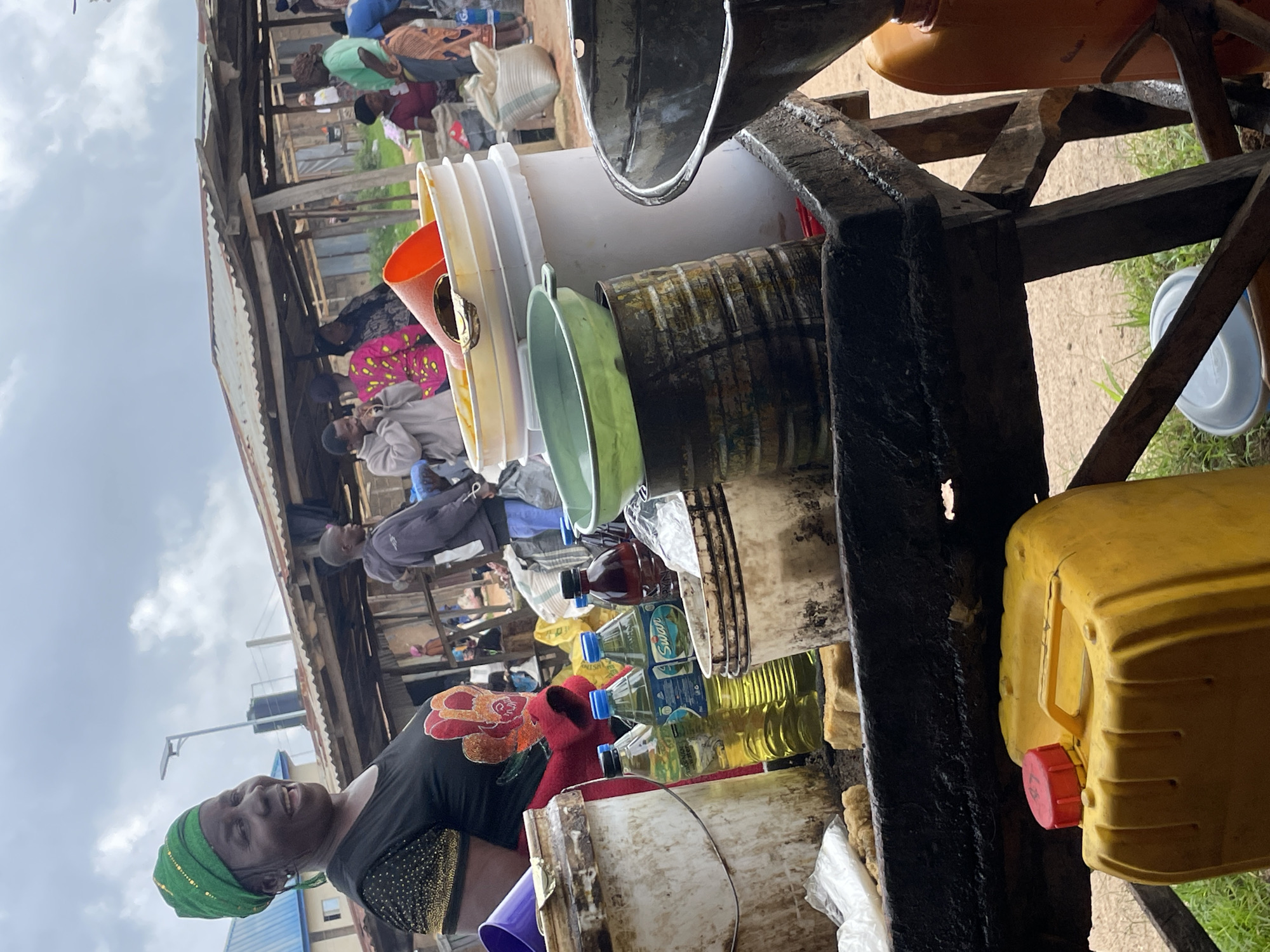 A woman wearing a green headwrap and a black shirt with gold embellishments smiles broadly at the camera in what appears to be an outdoor market. She is positioned in the foreground, with a wooden table laden with various containers and bottles in front of her. The table holds large plastic buckets, several bottles of yellow liquid, a dark red bottle, and metal containers. In the background, market stalls with corrugated metal roofs are visible, with other people milling about. The scene suggests a bustling marketplace environment, likely in a rural or semi-urban setting in Wang, Nigeria, given the presence of the woman and the style of the market stalls. The lighting suggests it is daytime, with an overcast sky.