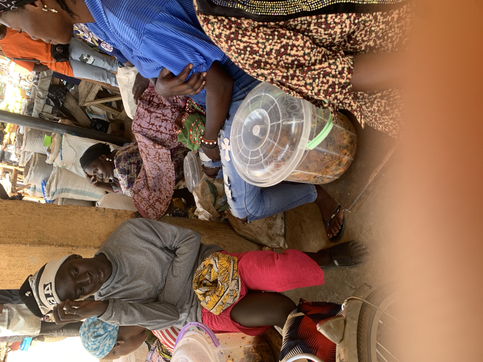 In Kafanchan B, Nigeria, a street market scene is captured. In the foreground, a young person wearing a gray long-sleeved shirt and a beanie hat with black and white stripes sits with their legs crossed, one foot bare and the other in a black sandal. A yellow and white patterned scarf is tied around their waist. To the left, a plastic container filled with food is visible, next to a bicycle wheel and frame. Further to the left, a person in a blue headscarf is partially visible, with their back to the camera.
In the mid-ground, two women are seated. One, on the left, wears a dark headscarf and a patterned dress, her right hand resting on her chin. The other woman, in the center, wears a pink and white patterned dress and a dark headscarf.
In the background, a person in an orange shirt and light blue jeans with a cat patch is standing. Bags of what appear to be grains or other market goods are stacked behind them, and wooden stalls are visible. The ground is dusty and uneven, consistent with an outdoor market environment. The overall lighting suggests it is daytime.