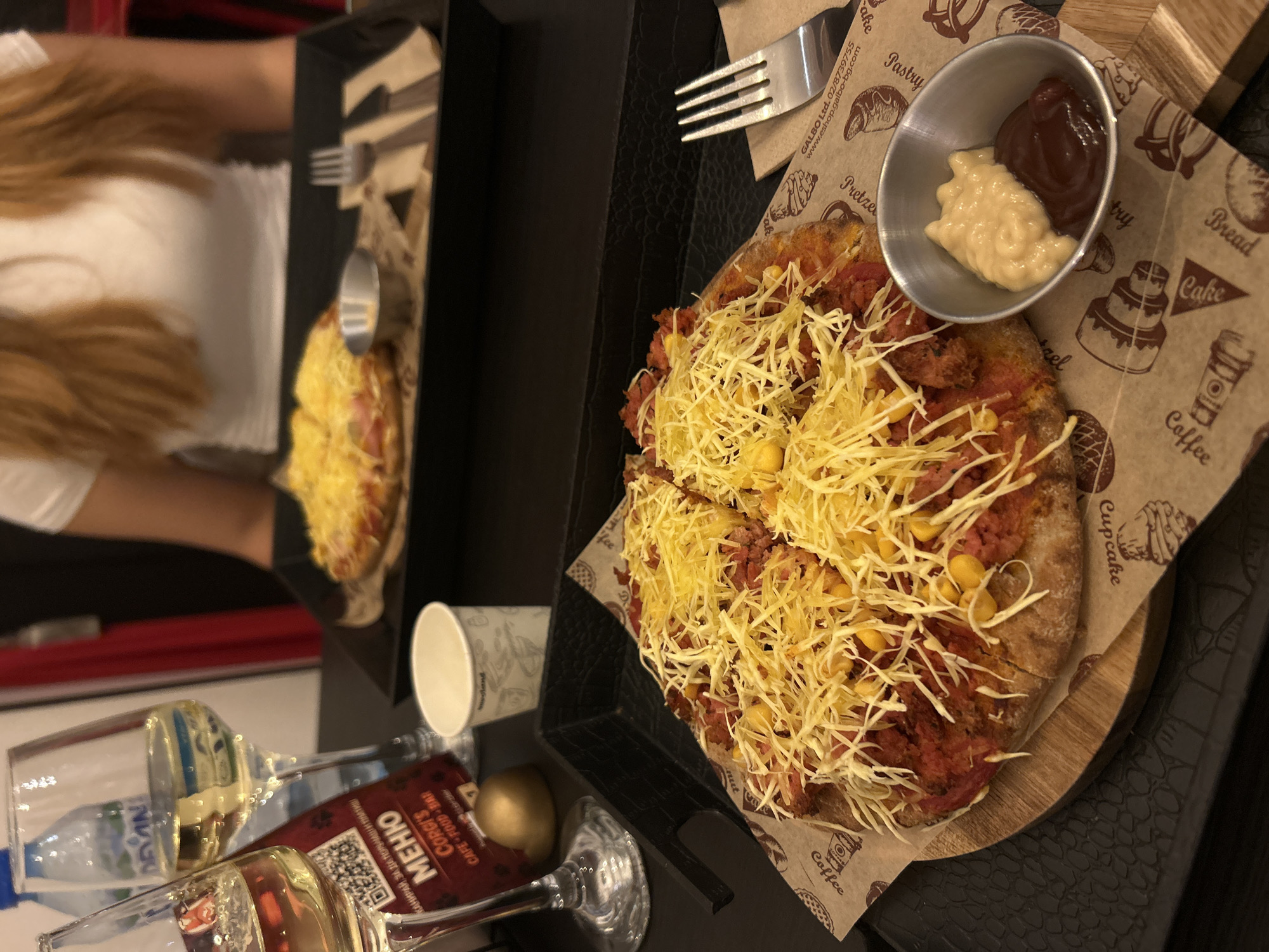 The image captures an overhead view of a casual dining setting, likely in a cafe or restaurant, where two people are enjoying food and drinks. The scene is dominated by a large, round pizza or flatbread, generously topped with shredded yellow cheese, corn kernels, and a reddish meat or sausage topping. This main dish is served on a wooden board placed on a black tray, over patterned paper featuring illustrations of bakery items like pretzels, bread, cake, and coffee. Adjacent to the pizza, a small metal cup holds two distinct sauces: a dark red sauce (likely ketchup or BBQ sauce) and a lighter, creamy sauce (possibly mayonnaise or a cheese dip).
To the left, a smaller, similar-looking flatbread is visible, accompanied by a fork, suggesting another serving. A person's arm and long, light brown hair are partially visible on the far left, indicating their presence at the table. On the bottom left, various drinks are arranged, including a plastic bottle of "Devin" brand water, and two wine glasses, one containing a red liquid. A white paper cup is also present.
The activity taking place is a meal or snack. Notable details include the patterned paper with text such as "GALBO Ltd.", "02/8739755", and "www.eshop.galbo-bg.com". A small card for "CORGI'S CAFE - FOOD - BAR" is visible, along with Cyrillic text "МЕНЮ" (Menu) and "КАНИРАЙ, ЗА ДА ПОРУЧАЦІИ ПЛАТИШ" (Scan to order and pay), confirming the location context of Sofia, Bulgaria, and hinting at a modern ordering system. The time of day is not explicitly indicated but appears to be indoors under artificial lighting. No specific emotions are discernible, but the setup suggests a relaxed dining experience.