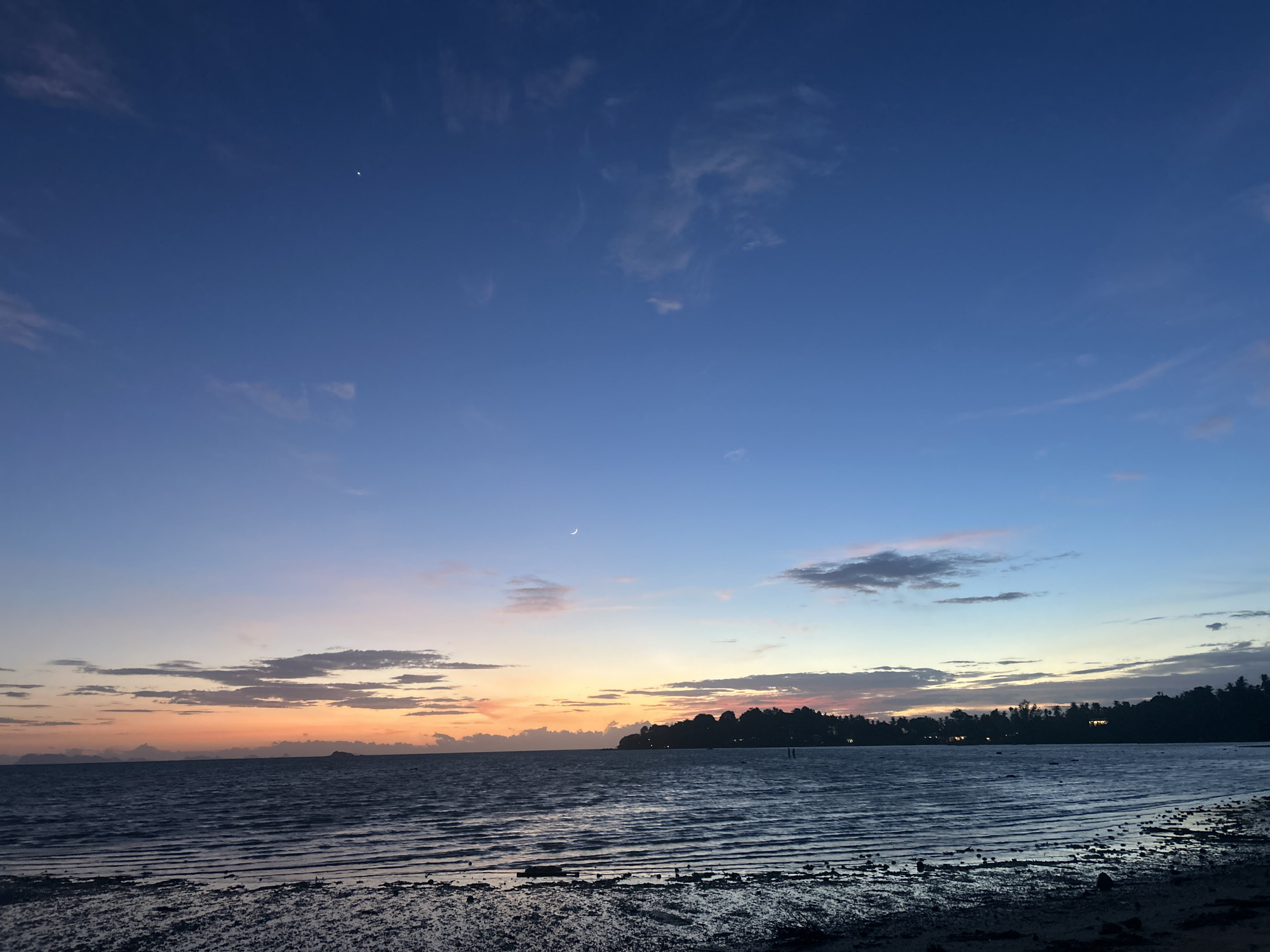This is a photograph of a twilight scene on the coast of Ko Pha-ngan District, Thailand. The sky transitions from a deep blue at the top to orange and pink hues near the horizon, with scattered clouds. A crescent moon is visible in the upper left quadrant of the sky. The sea is dark and choppy, with visible ripples. In the foreground, the shore is composed of wet sand and scattered rocks, indicating a low tide. Silhouetted against the horizon is a tree line with scattered lights, suggesting a settlement or buildings along the coast. The overall impression is one of a serene coastal sunset.