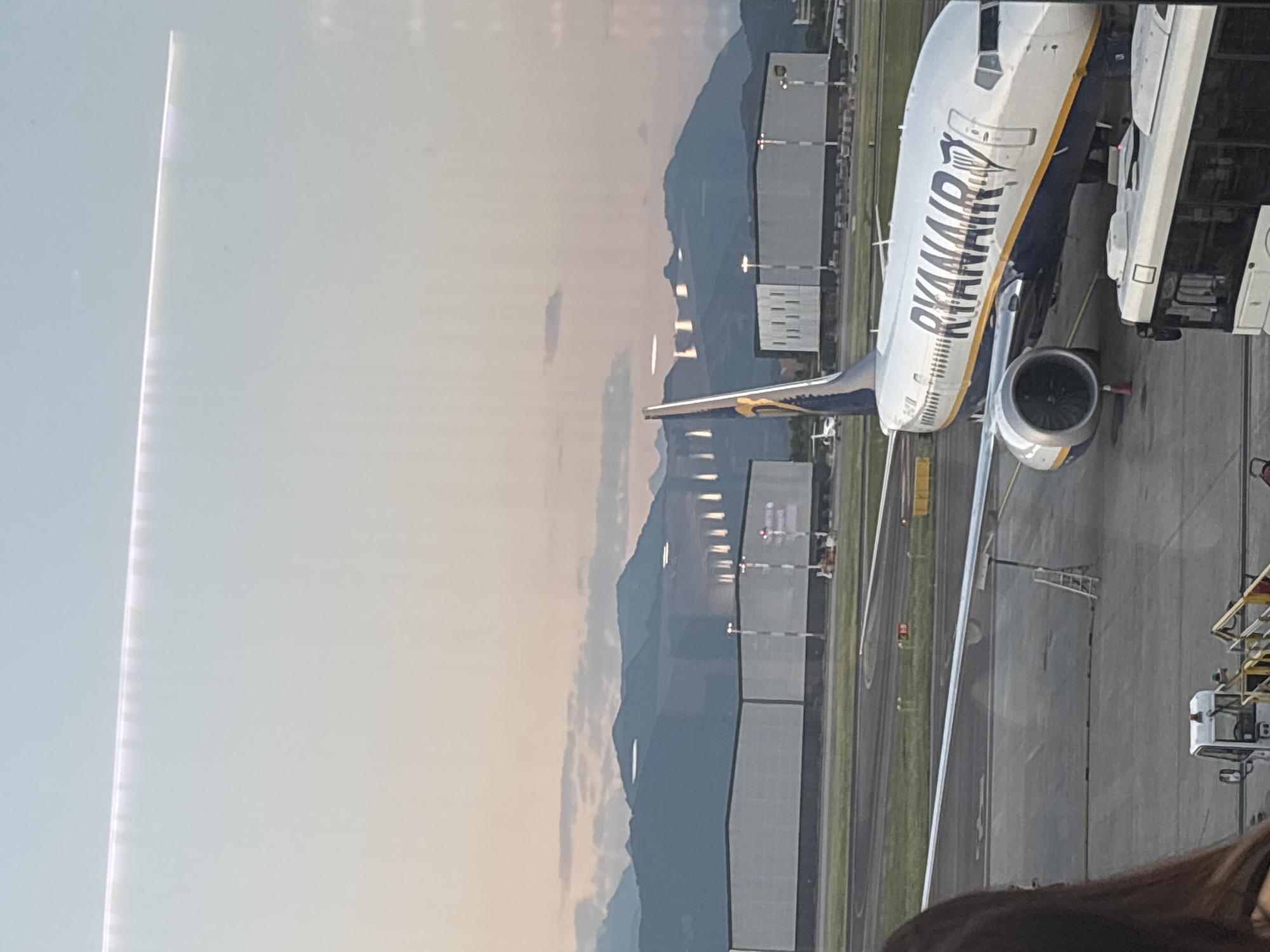 The image captures a Ryanair airplane parked on the tarmac at Orio al Serio International Airport (BGY) in Orio al Serio, Italy. The aircraft, predominantly white with blue and yellow accents, displays the "RYANAIR" logo prominently along its fuselage. Its engines are visible, and a set of stairs is positioned near the front of the plane, suggesting it is either preparing for departure or has recently arrived. In the foreground, a portion of a white bus is visible, indicating passenger transport operations. Beyond the airplane, several large, grey hangar-like structures stand adjacent to the runway. In the background, a range of mountains forms the horizon under a sky transitioning from pale blue at the top to a soft pink and orange hue near the horizon, suggesting either sunrise or sunset. The scene is set in an airport environment with typical tarmac infrastructure, including lighting and markings.