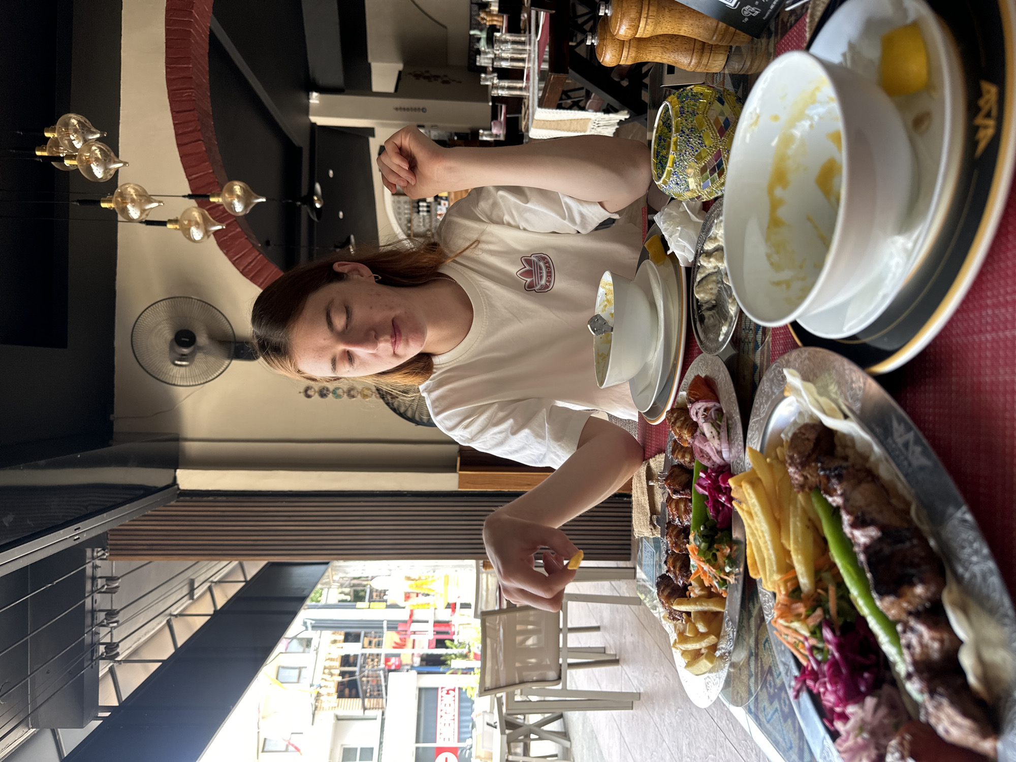 The image shows a person, presumably female with light brown hair and wearing a white Adidas t-shirt, seated at a table laden with food in a restaurant. The person is holding a french fry.
The food on the table includes what appears to be a mixed grill, a bowl of soup, and various side dishes. The table is covered with a red cloth and a second serving platter.
Behind the person is an interior view of the restaurant, featuring a black and white patterned wall, a fan, and distinctive lighting fixtures. Beyond the restaurant's window, an external cityscape of Baan Chaweng, Thailand is visible, with buildings, street signs, and trees under daylight conditions. Tables and chairs can be seen outside the establishment.