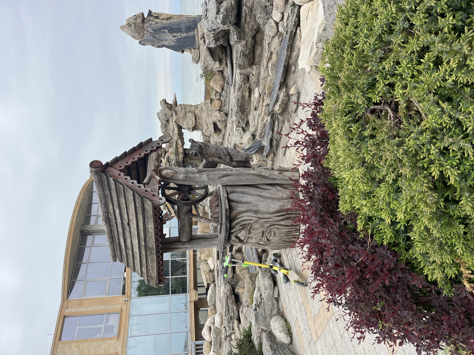 The image shows a decorative well structure constructed of wood, complete with a wheel and a roof. The well is situated amidst a rock garden and surrounded by shrubs, including both green and burgundy varieties. Adjacent to the rock garden is a modern building with large, arched windows. A scooter lies near the well. In the background, a rocky coastline is visible under an overcast sky. The scene is located in the city of Kaspiysk, Russia.