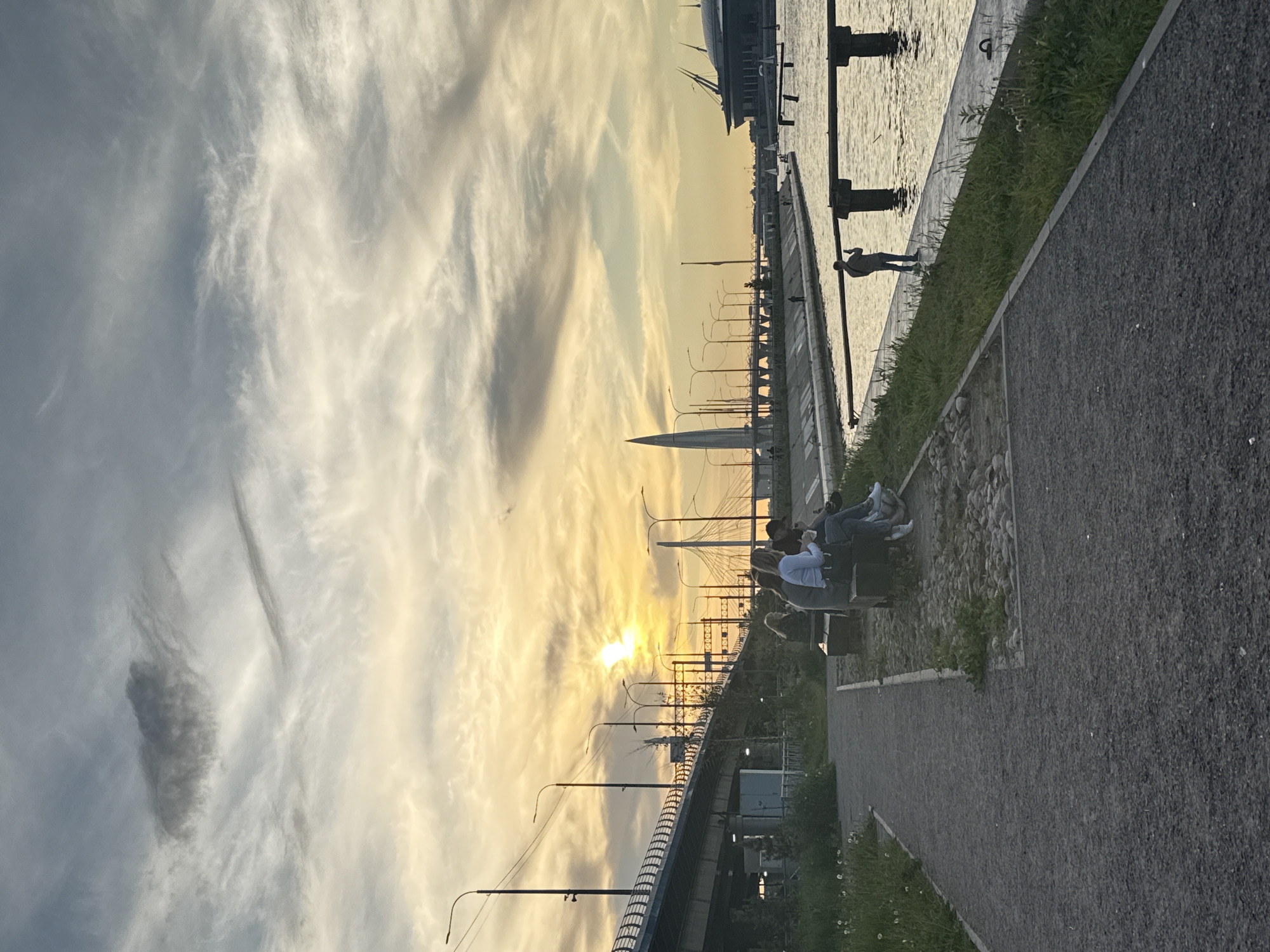 The image, taken during sunset or late afternoon, depicts a serene waterfront promenade in Saint Petersburg, Russia. The sun, low on the horizon, casts a warm, golden glow across the entire scene, particularly illuminating the dramatic, wispy clouds in the sky.
In the mid-ground, three individuals are seated on a bench, facing away from the camera, seemingly engrossed in the view of the water and the setting sun. Further along the paved pathway, another person stands near the water's edge. The foreground features a pathway, a strip of green grass, and some scattered rocks.
To the left, a body of water reflects the golden light of the sky, with several dark bollards or mooring posts visible along its edge. The background is dominated by the silhouetted skyline of a city, featuring various buildings, numerous streetlights lining a road or bridge structure, and a prominent, modern cable-stayed bridge, likely part of the city's infrastructure (such as the Western High-Speed Diameter bridge). The sun is partially obscured by the clouds and the bridge's framework.
The overall atmosphere is peaceful and reflective, with the warm light enhancing the natural beauty of the clouds and the urban landscape. There is no visible text in the image.
