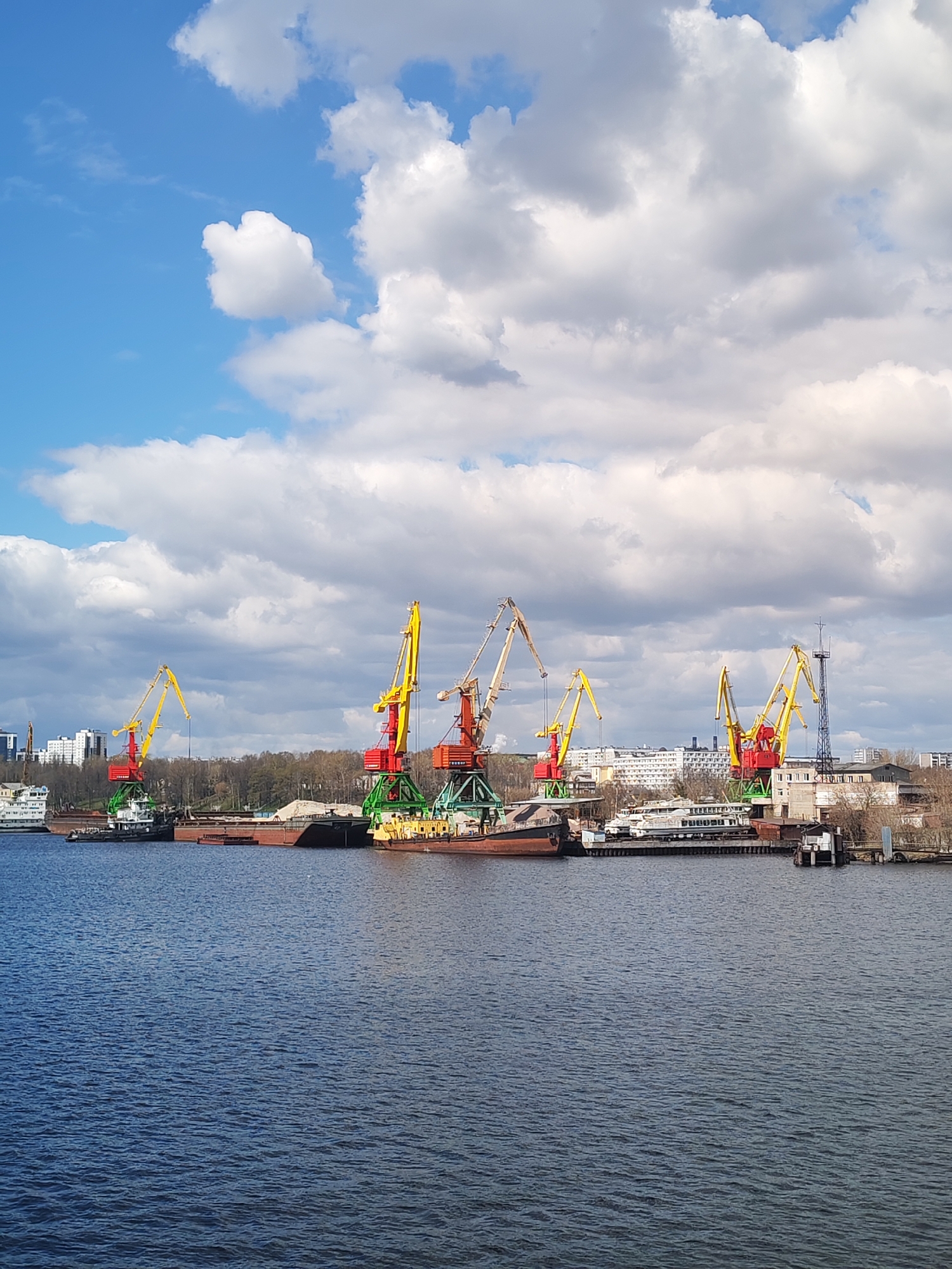 This image depicts an industrial river port in Cherepovets, Russia, under a partially cloudy sky. In the foreground, a wide expanse of dark blue water stretches across the frame, showing ripples on its surface. The midground is dominated by a series of large port cranes, most prominently colored in bright yellow and red, indicating their industrial purpose. At least five such cranes are visible along the riverbank.
Dotted along the shore beneath the cranes are several barges, some appearing to be loaded with bulk materials like sand or gravel. A white passenger boat or ferry is also docked further down the river, alongside other smaller vessels and industrial structures. The background features a mix of industrial buildings and structures, interspersed with trees, and in the distance, more urban buildings are visible, suggesting a city's proximity to the port.
The sky above is a vibrant blue, with numerous large, fluffy white cumulus clouds scattered across it, indicating a clear, possibly breezy, daytime. The overall impression is one of an active working port. No people are visible in the scene, and there is no discernible text within the image. The setting conveys a sense of industrial activity and cargo transportation along the river.