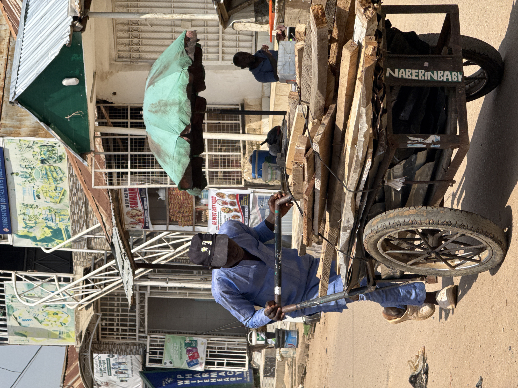 A man in a blue tunic and black baseball cap pushes a wooden cart loaded with lumber down a dusty, unpaved street in Abuja, Nigeria. The cart has two large spoked wheels and a metal frame. He is wearing white shoes. In the background, a green umbrella shades a vendor. Buildings with corrugated metal roofs and signs in English line the street. One sign reads "WHITEHEART PHARMACY" and another displays food images and the text "DE BEST FAST FOOD SHAWARMA". The man appears to be moving the lumber, possibly for construction or sale. The scene suggests a busy marketplace or commercial area in the city.