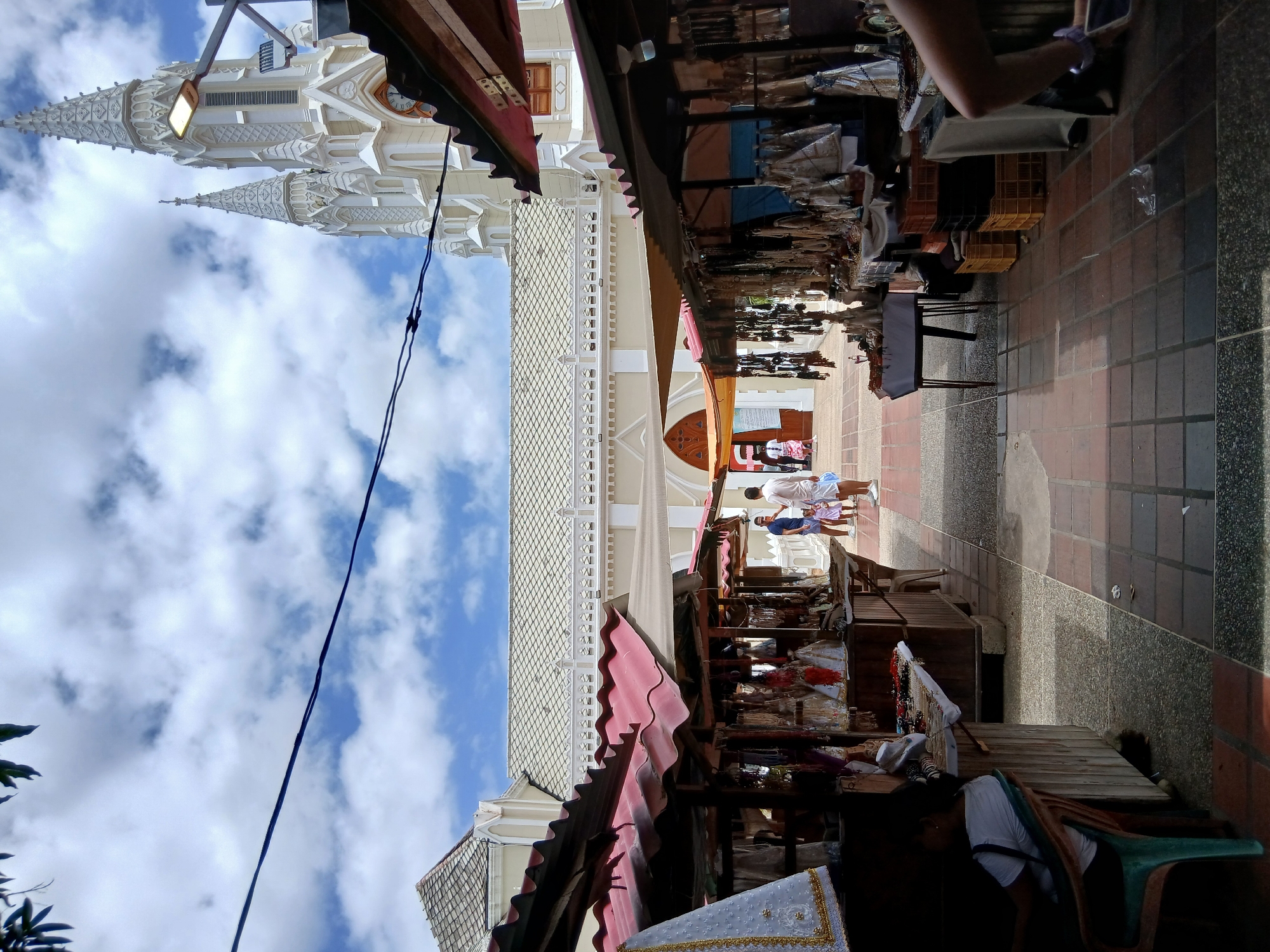 The image captures a street scene in Altagracia, Venezuela, flanked by market stalls on one side and a large white church on the other. A paved walkway runs between the stalls and the church, with a section of red and gray brickwork. People are visible in the distance along the walkway. The church's architectural details, including its towers and ornate trim, are visible against a partly cloudy sky. The market stalls have red awnings and display a variety of merchandise. A person's arm is visible in the upper-right corner, holding a phone.