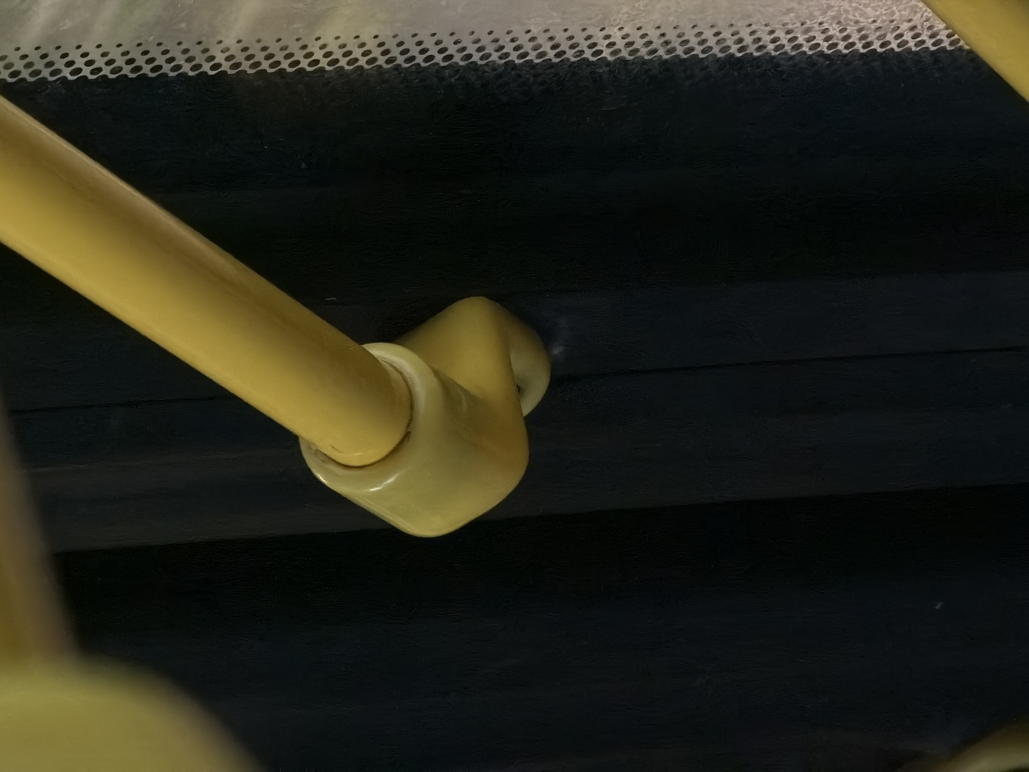 The image captures a close-up view of a yellow handrail on a dark background, likely inside a public transport vehicle in Butovo, Russia. The handrail appears to be made of sturdy plastic and is designed with a curved section and a circular bracket, suggesting it's part of a larger support system. Above the handrail, a strip of white material with a pattern of small, dark dots is visible, possibly a decorative element or a way to improve grip on the vehicle's interior surface, perhaps near a window. The overall lighting suggests an interior setting, possibly daytime given the potential for ambient light from a window, though the darkness of the background dominates the scene. No people are visible, and no specific event is taking place; the image focuses solely on the architectural details of the vehicle's interior. There is no visible text in the image.