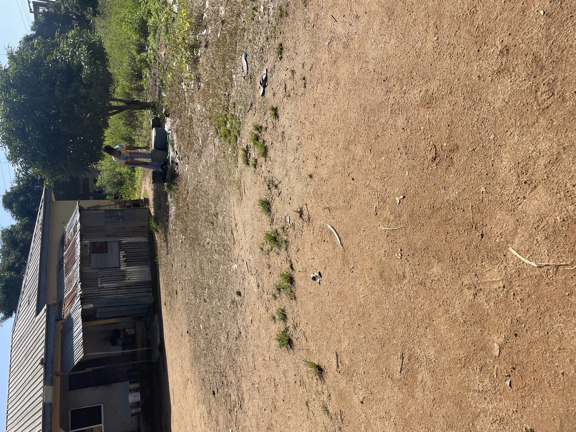 In the city of Jos, Nigeria, a young person, likely a girl wearing a white t-shirt and patterned trousers, stands by a concrete well, drawing water. She is positioned under a tree with lush green foliage. To the left of the well, several large blue containers are visible. In the background, a simple, single-story house is situated, constructed with a corrugated metal roof and partially covered by a corrugated metal awning in front of a section that appears to be a makeshift structure. Buckets are placed near the entrance of the house. The ground is a mixture of dry earth, scattered dry grass, and some debris. A pair of flip-flops lies on the ground a short distance from the well. The overall environment suggests a rural or semi-rural setting within Jos.