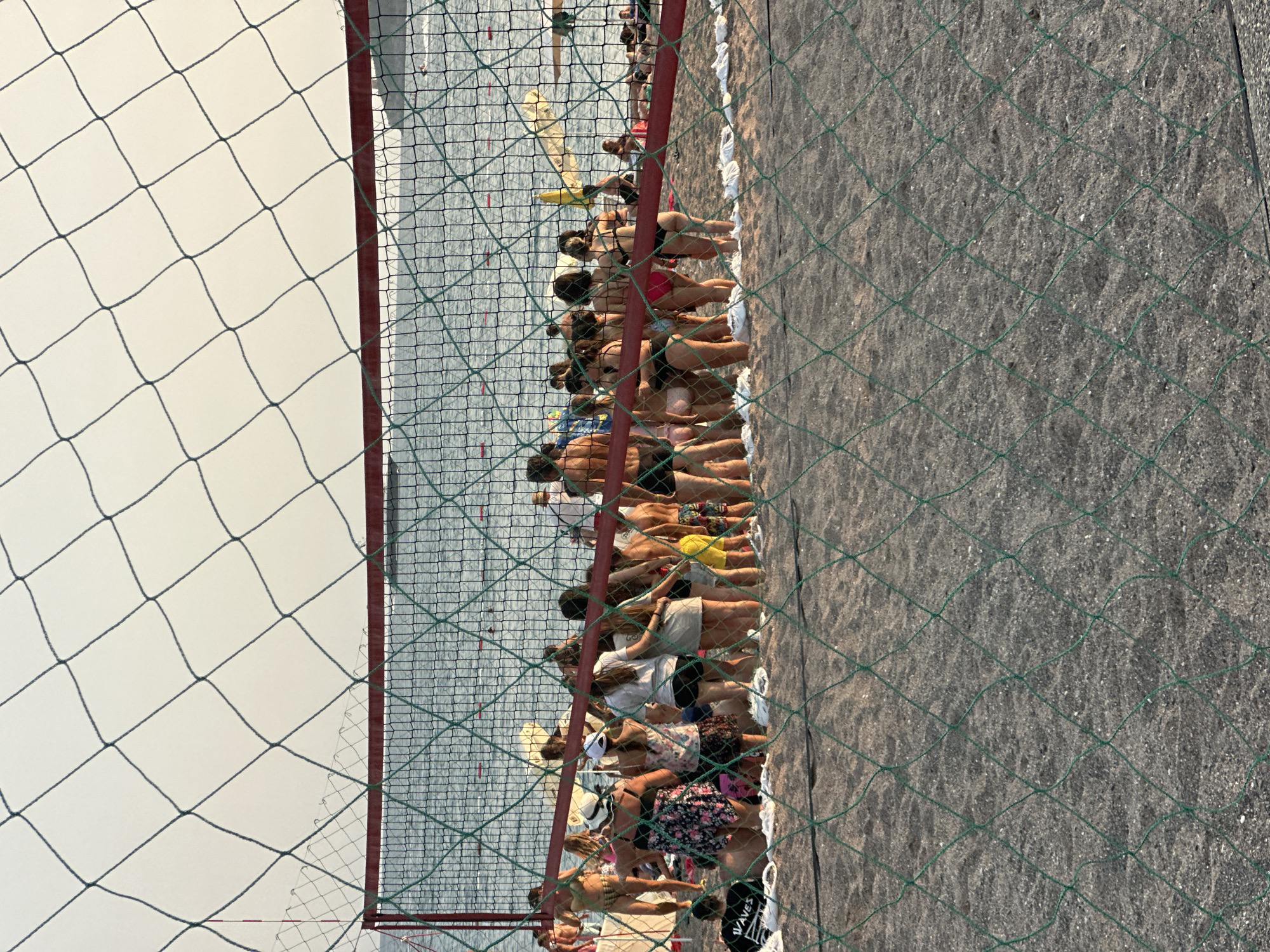 In Boreti, Montenegro, a group of individuals are gathered on a sandy beach in front of a volleyball net. The net, with its green mesh and red top frame, is in the foreground and partially obscures the scene. The water is visible behind the net, with gentle waves and what appear to be buoys in the distance. Several people are standing in a cluster, facing away from the camera, some in swimwear, suggesting a casual social gathering. The sand is light in color, and the overall lighting indicates it is either early morning or late afternoon. The environment suggests a recreational area, likely a beach resort.
