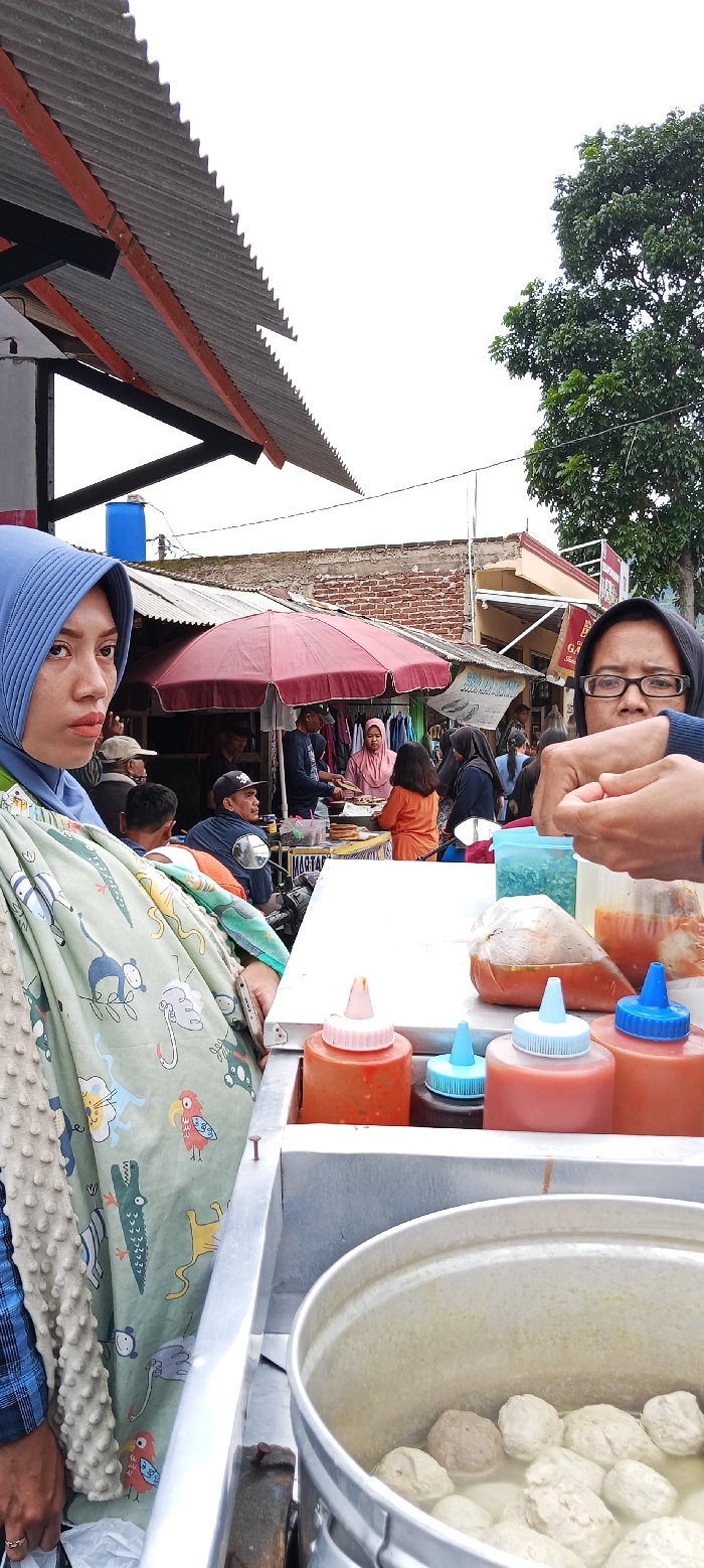 The scene takes place in a market in Kecamatan Kutawaringin, Indonesia, at latitude -6.973 and longitude 107.52. A woman wearing a blue headscarf and a green and white patterned shawl stands at a food stall, looking at the camera. In front of her is a metal counter with a pot of meatball soup. The pot is surrounded by containers filled with chili sauces and other condiments. Several other people are visible in the background, some are buying food, others are walking by. There are buildings and trees in the background. The stall is covered by a corrugated metal roof. The scene appears to be on a busy street, with a lot of pedestrian activity. The woman on the left is holding a bag in her left hand. She is looking at the camera. There is a man standing behind her, holding his phone. He is wearing a white shirt and a blue cap.